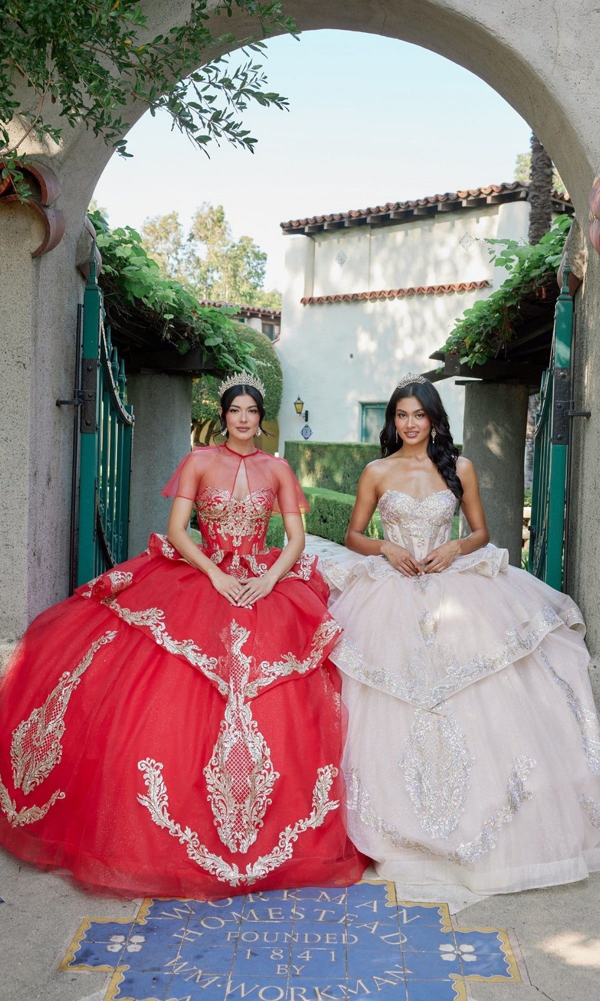 Two women stand side by side in ornate ball gowns—one wearing the Quinceanera Dress JT1511H By Juliet, a striking red gown adorned with gold applique embroidery, and the other in a graceful white strapless ballgown—posing under an arched stone entrance with greenery and a tiled floor visible.