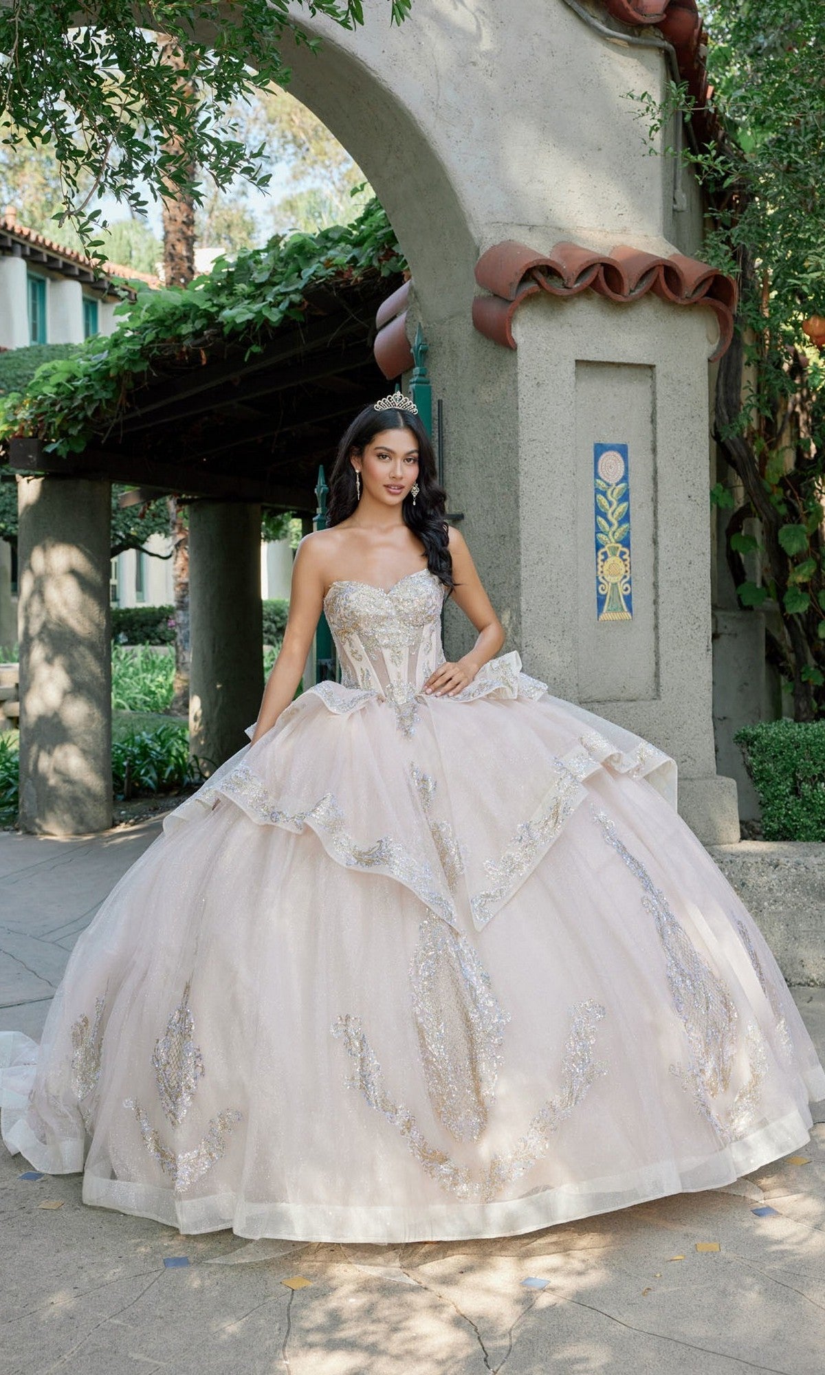 A woman wearing the Quinceanera Dress JT1511H By Juliet in champagne, a strapless ballgown featuring gold applique embroidery, stands outdoors beside a stucco archway with lush greenery in the background.