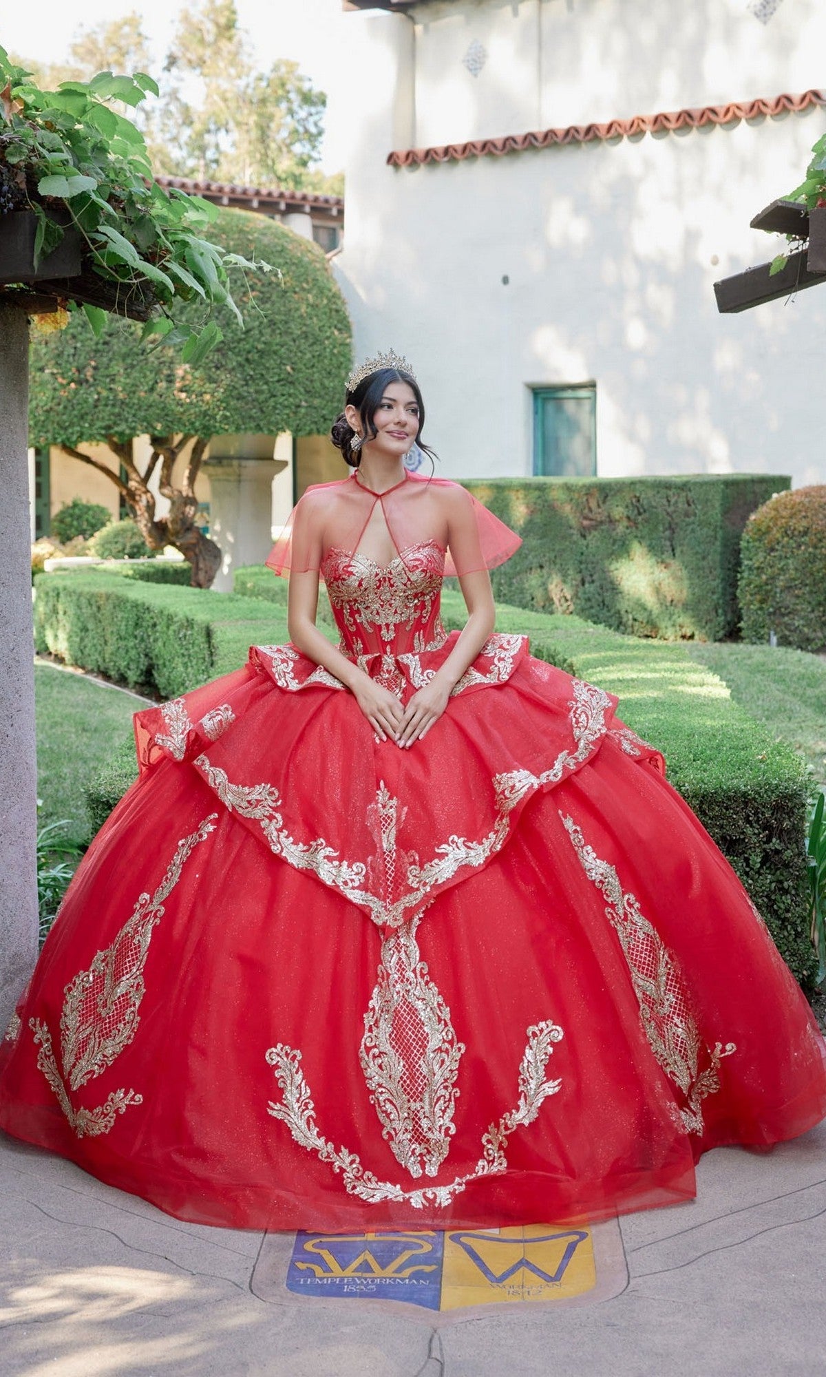A young woman stands outdoors in a manicured garden, hands clasped in front of her and framed by greenery and a white building with a tiled roof in the background. She is wearing the Quinceanera Dress JT1511H By Juliet in Red/Gold, featuring ornate gold applique embroidery and accessorized with a tiara.