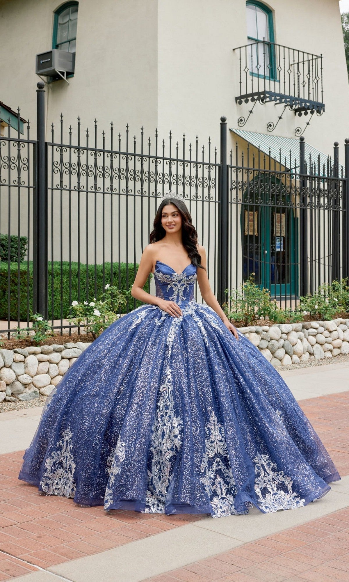 A young woman wearing the Quinceanera Dress JT1512H By Juliet in navy blue stands outside in front of a wrought iron fence and a building with green trim. The sparkling glitter ballgown skirt and elegant corset bodice of her gown add to her graceful presence.