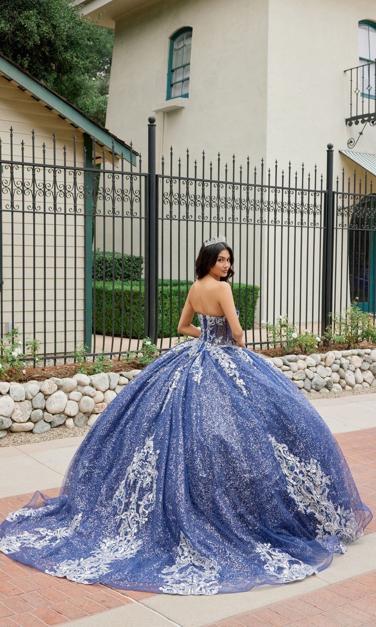 Wearing the Quinceanera Dress JT1512H by Juliet, a woman stands outside in front of a building and black iron fence. The strapless, blue and silver glitter ballgown features a corset bodice as she looks over her shoulder.