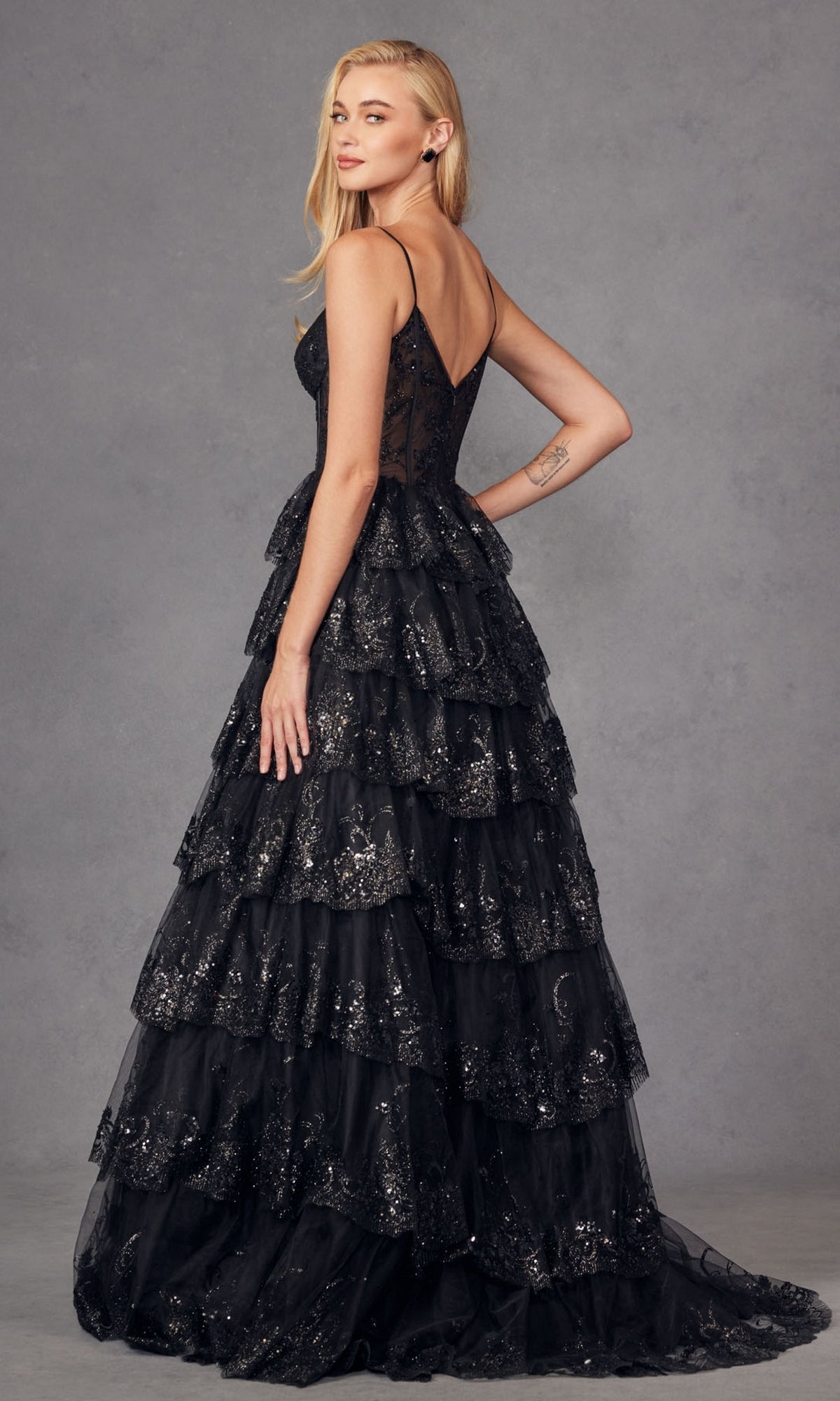 A woman poses against a plain gray background, looking over her shoulder in the Juliet JT2454K Long Ruffled A-Line Prom Ball Gown—a sleeveless black dress with tiered ruffles and glitter details.
