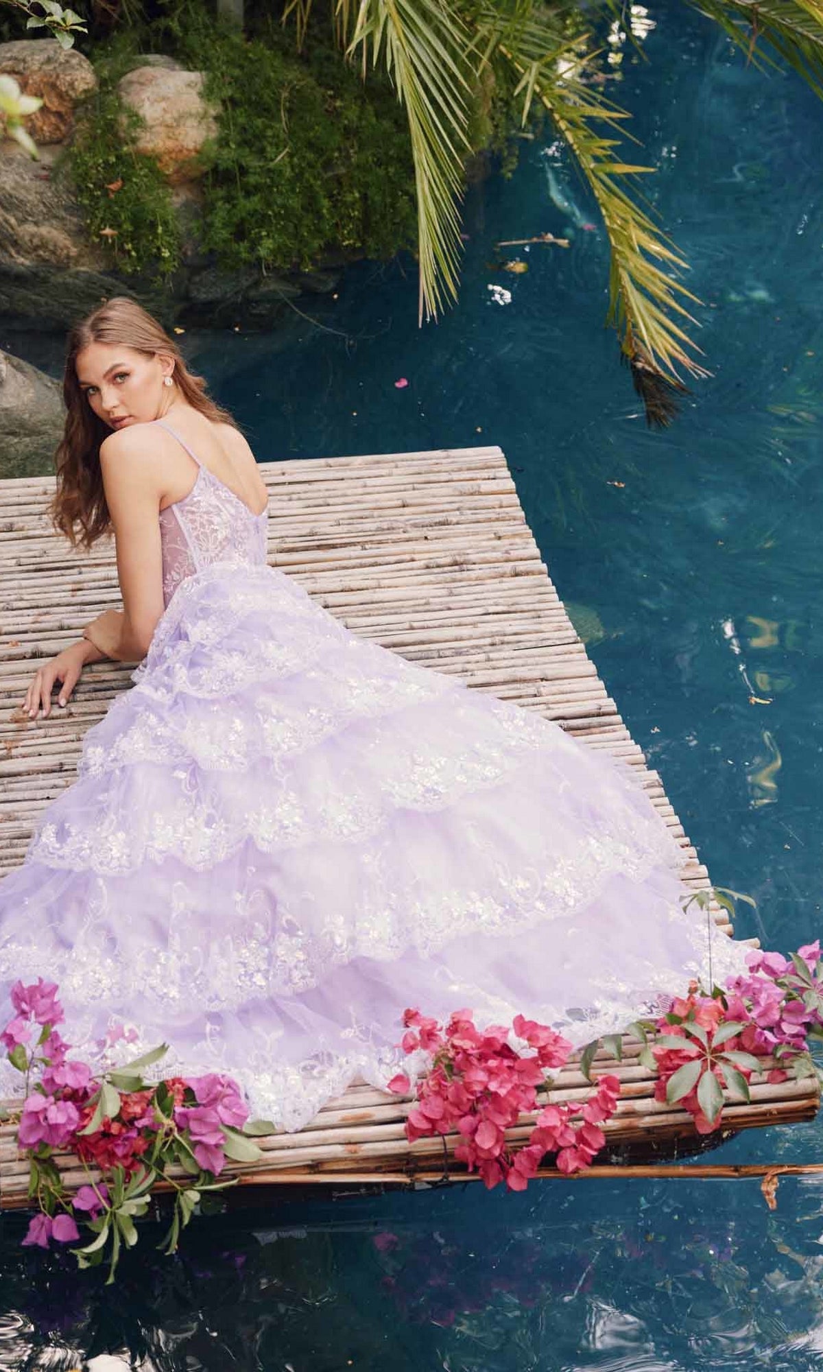 A woman in the Juliet JT2454K Long Ruffled A-Line Prom Ball Gown, lavender, sits on a wooden dock by blue water, surrounded by pink flowers and green foliage.
