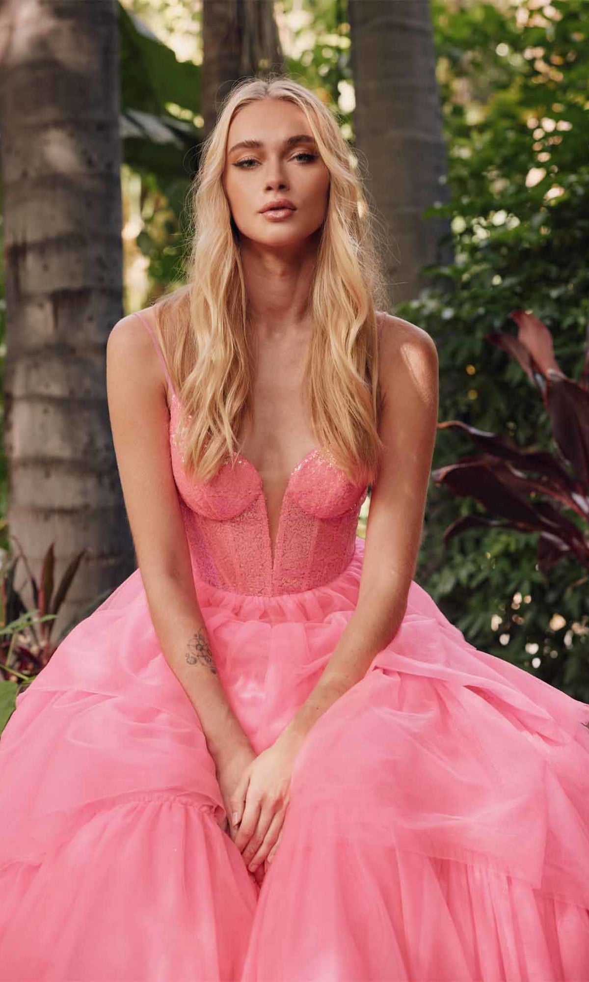 A woman with long blonde hair sits outdoors among green foliage, wearing the strapless Long Prom Dress JT2457H by Juliet in bright fuchsia.