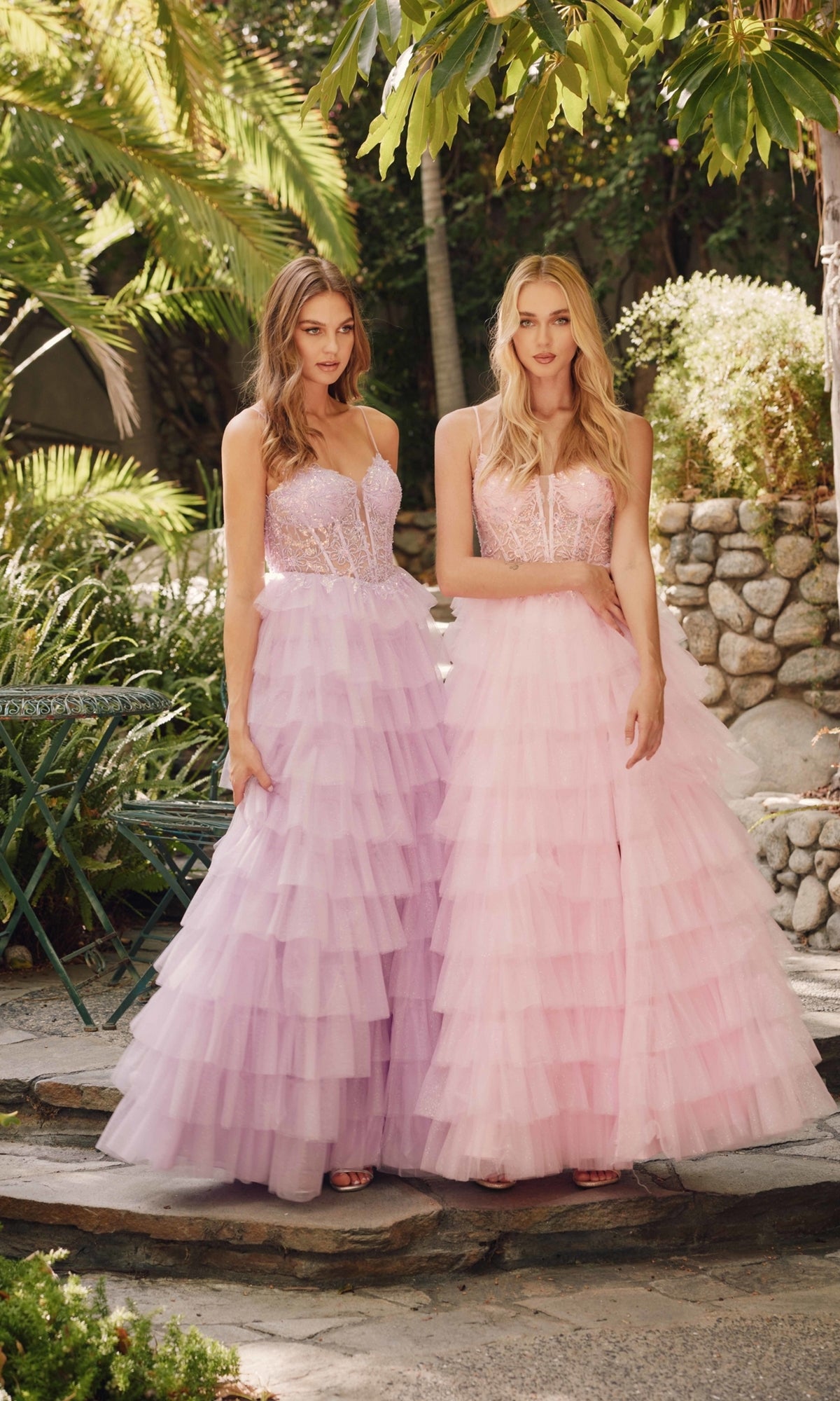 Two women stand outdoors in ruffled glitter tulle prom dresses, style Juliet JT2458A, in pastel pink and lavender. Amid greenery and stonework, the gowns’ tiered layers create a dreamy, elegant effect.
