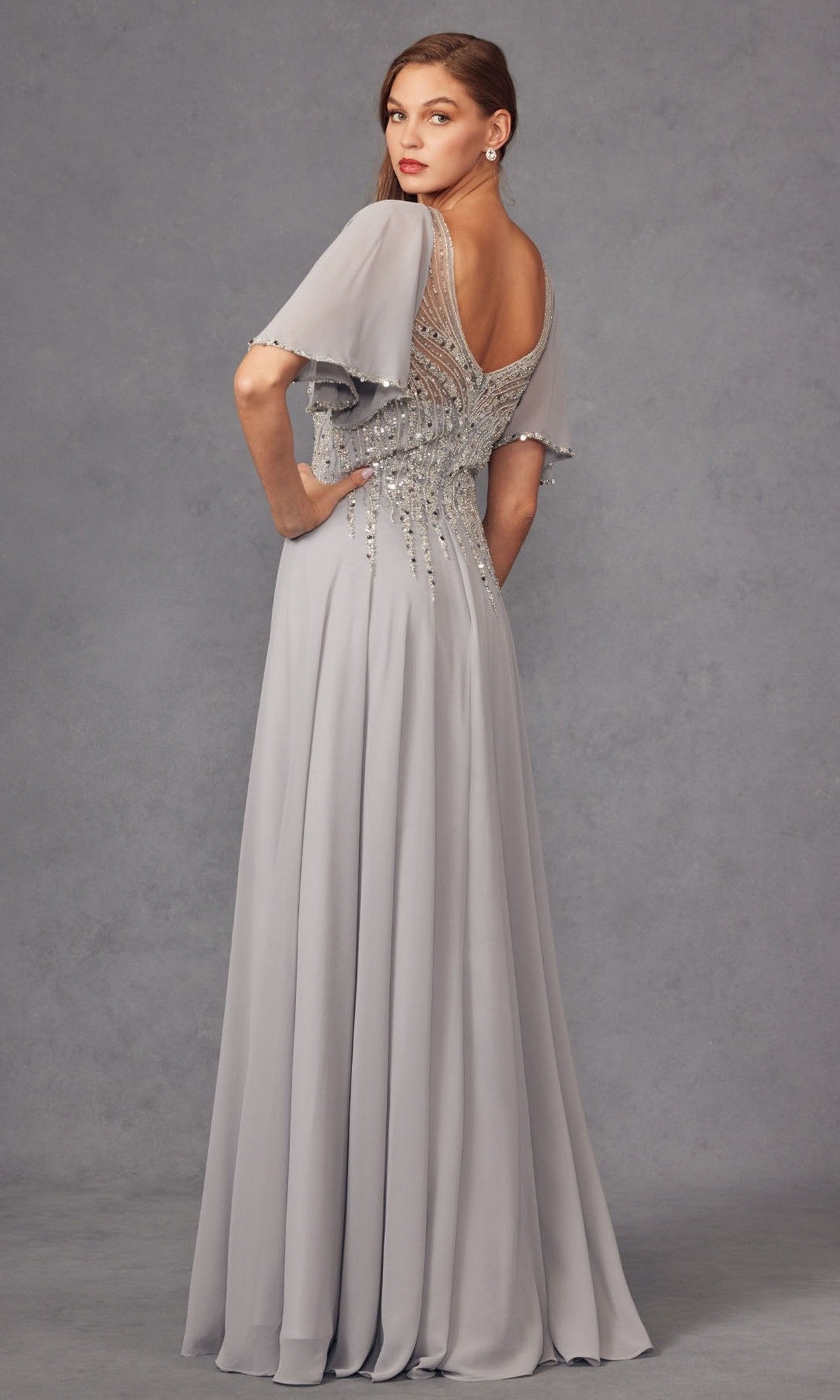 A woman stands against a plain gray backdrop wearing the Juliet JTM14F, a long formal silver gown featuring beaded detailing, short flutter sleeves, and a low back—an elegant choice reminiscent of a sophisticated mother of the bride dress.