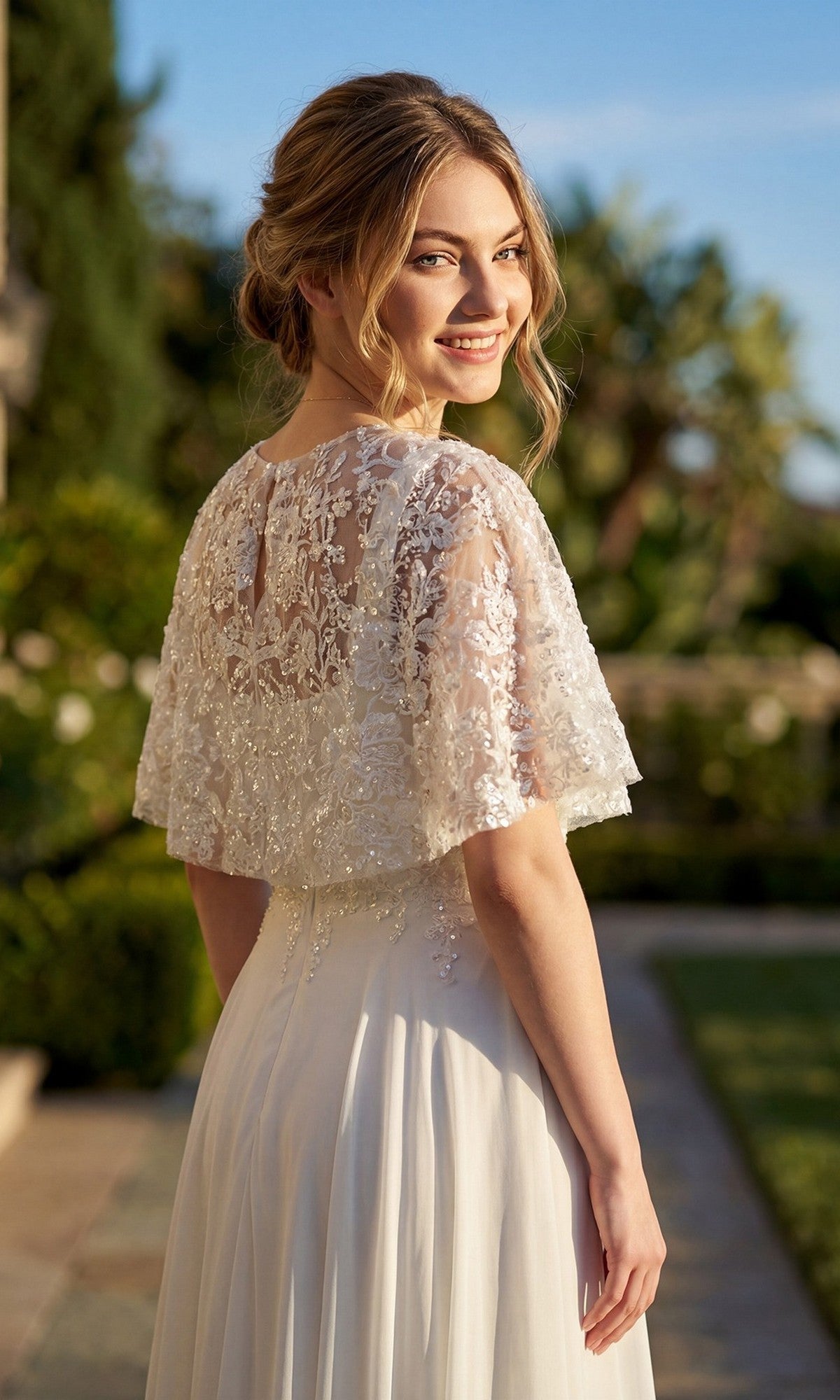 A woman wearing the Juliet JTM21LW long formal dress stands outdoors, smiling over her shoulder with greenery and sunlight in the background.