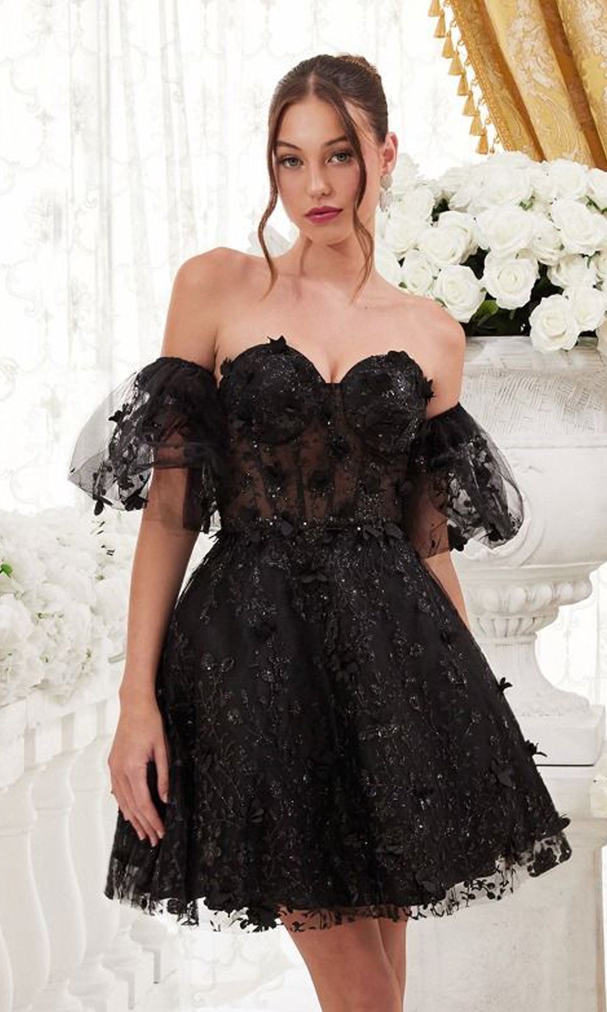 A woman wears the Puff-Sleeve Short Strapless Homecoming Dress KV1089, featuring black floral embellishments and sheer puff sleeves, while standing indoors near white floral arrangements and gold drapery.