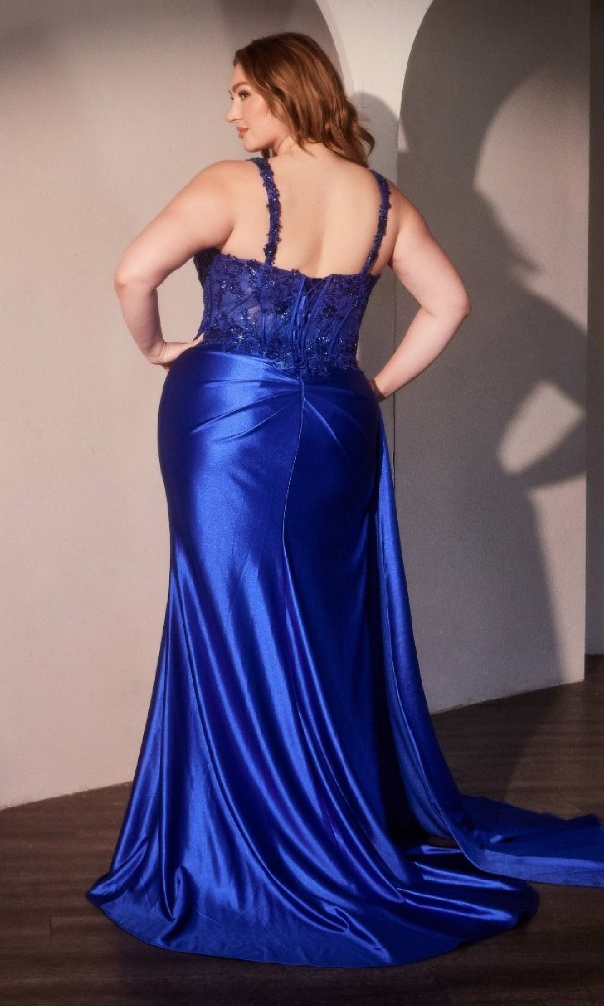 A woman stands with her back to the camera wearing the Side-Sash Plus-Size Prom Gown: Ladivine KV1125C, a floor-length evening dress in royal blue with lace and satin details.