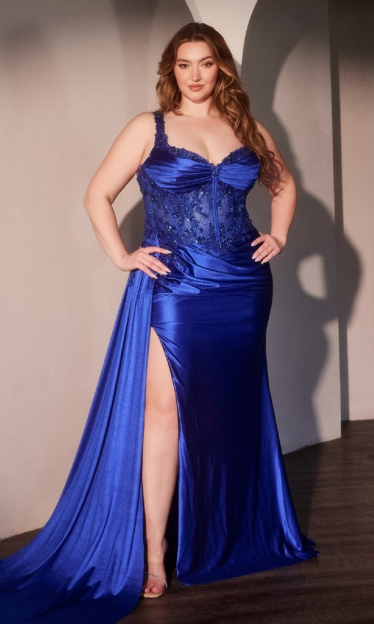 A woman wearing the Side-Sash Plus-Size Prom Gown (Ladivine KV1125C), a royal blue, floor-length satin dress with lace accents and a thigh-high slit, stands on an indoor wooden floor.