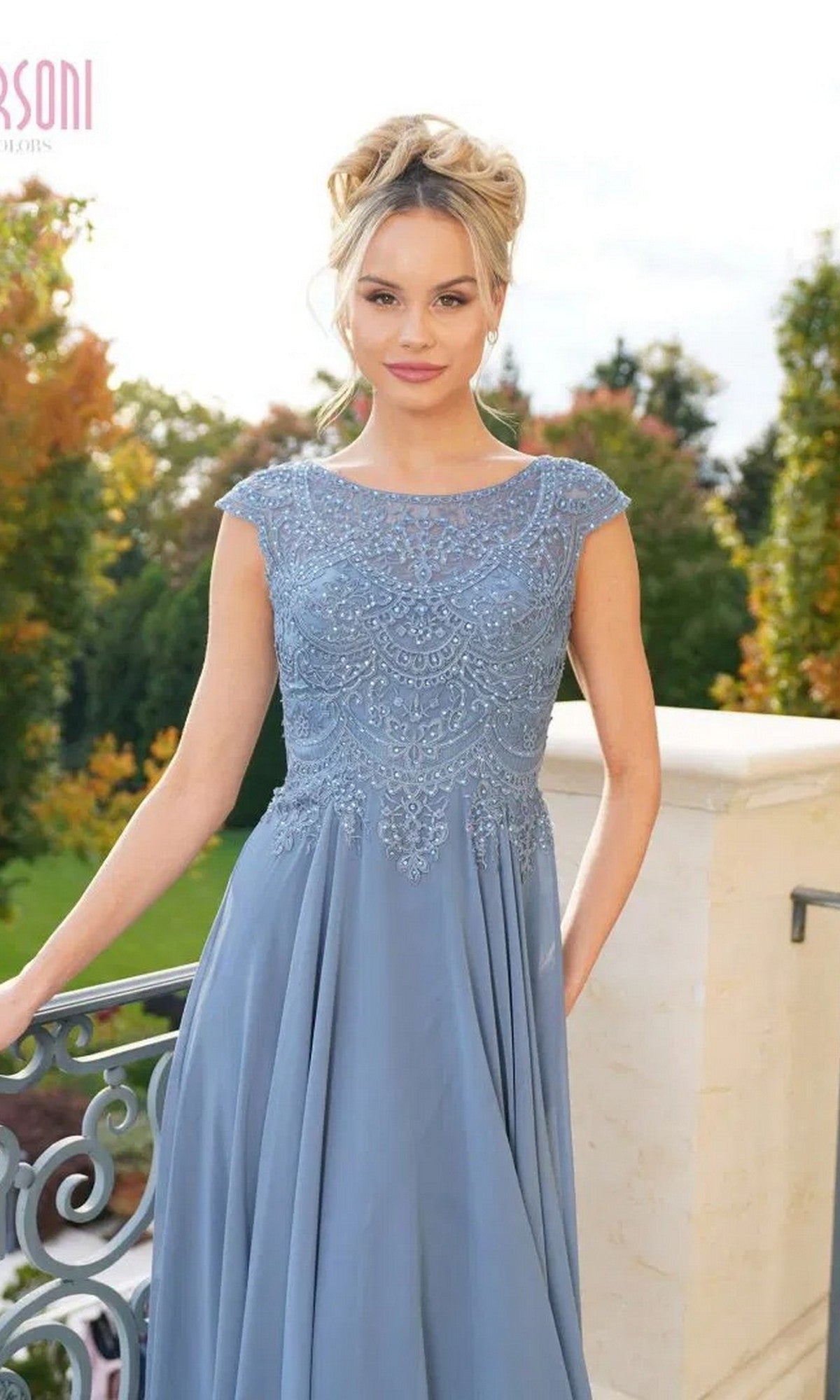 A woman wears the Marsoni M238 long formal dress—light blue, floor-length, with an embroidered bodice—standing outdoors by a railing with green trees behind her.