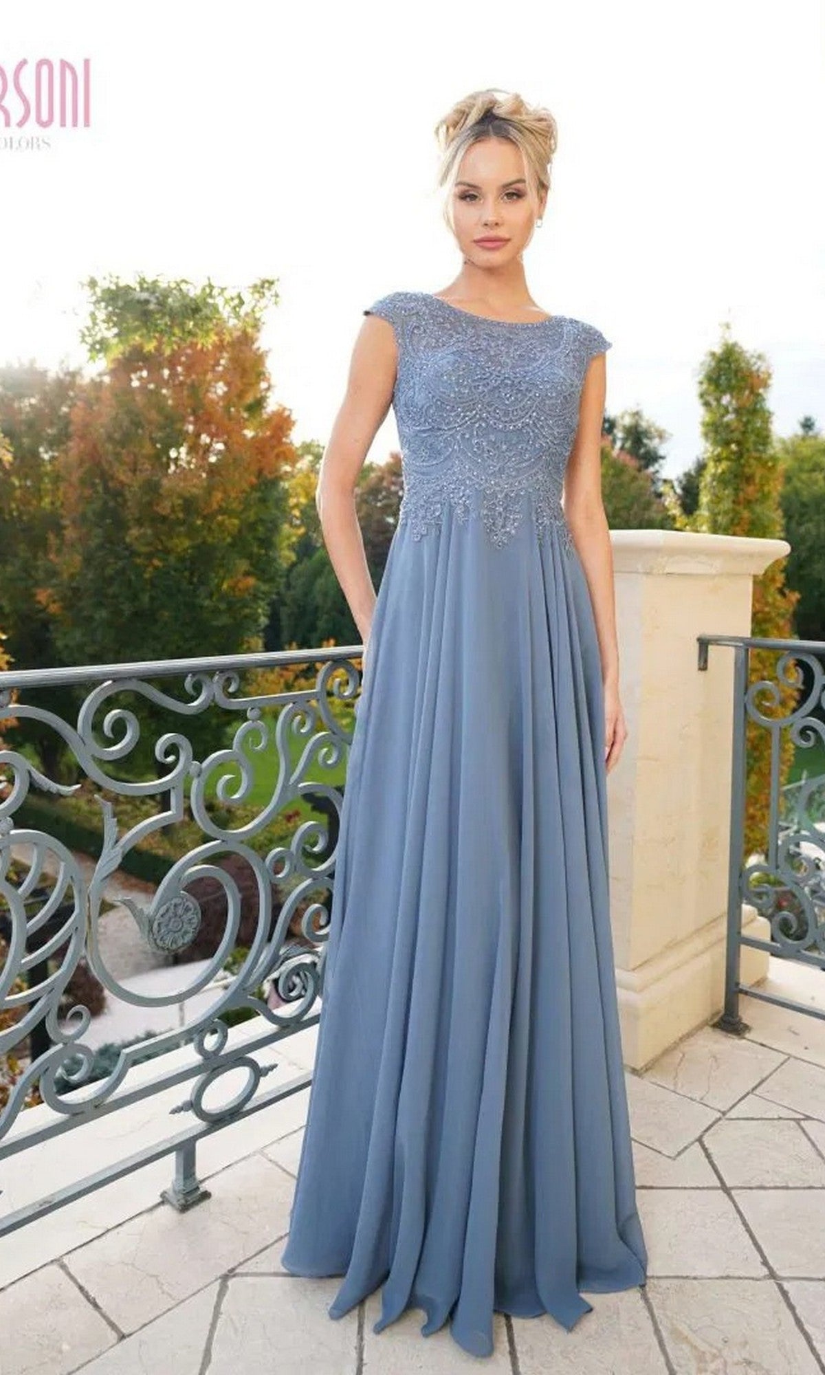A woman stands on a balcony in the Marsoni M238, a floor-length light blue formal dress with an embroidered bodice and flowing chiffon skirt, surrounded by greenery.
