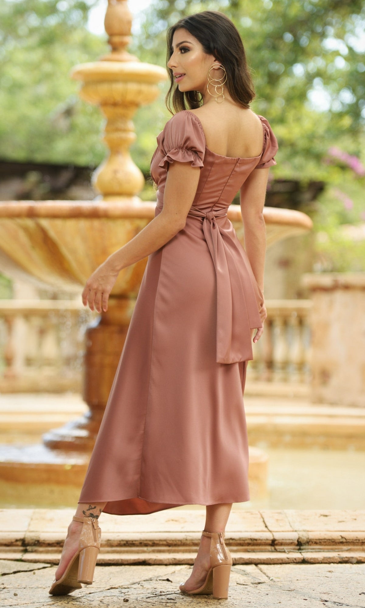 A woman in the Velvi Tea-Length Wedding-Guest Dress Maldives and clear heels stands by a stone fountain outdoors, glancing over her shoulder.