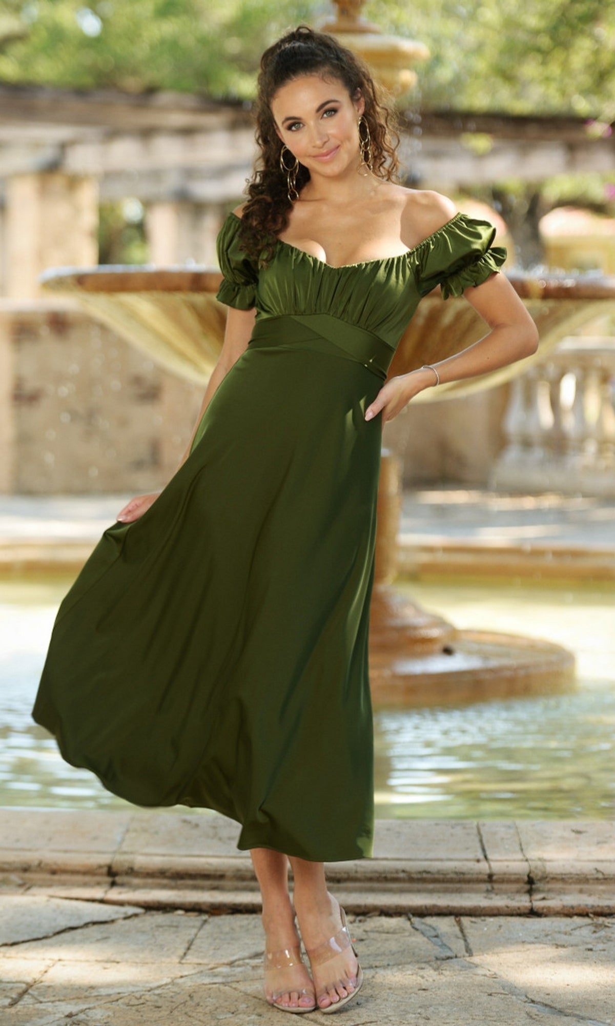 A woman in the Velvi Tea-Length Wedding-Guest Dress Maldives stands before a stone fountain, holding her dress and smiling at the camera.