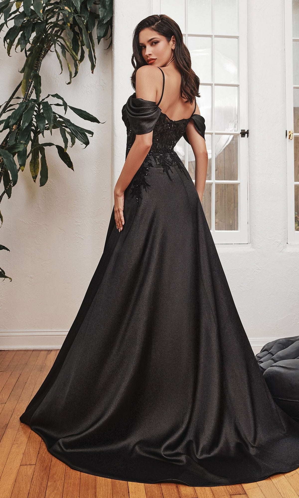 A woman stands on a wooden floor, facing away, wearing the Cold-Shoulder Long Satin A-Line Prom Dress OC012, with large windows and a leafy plant in the background.