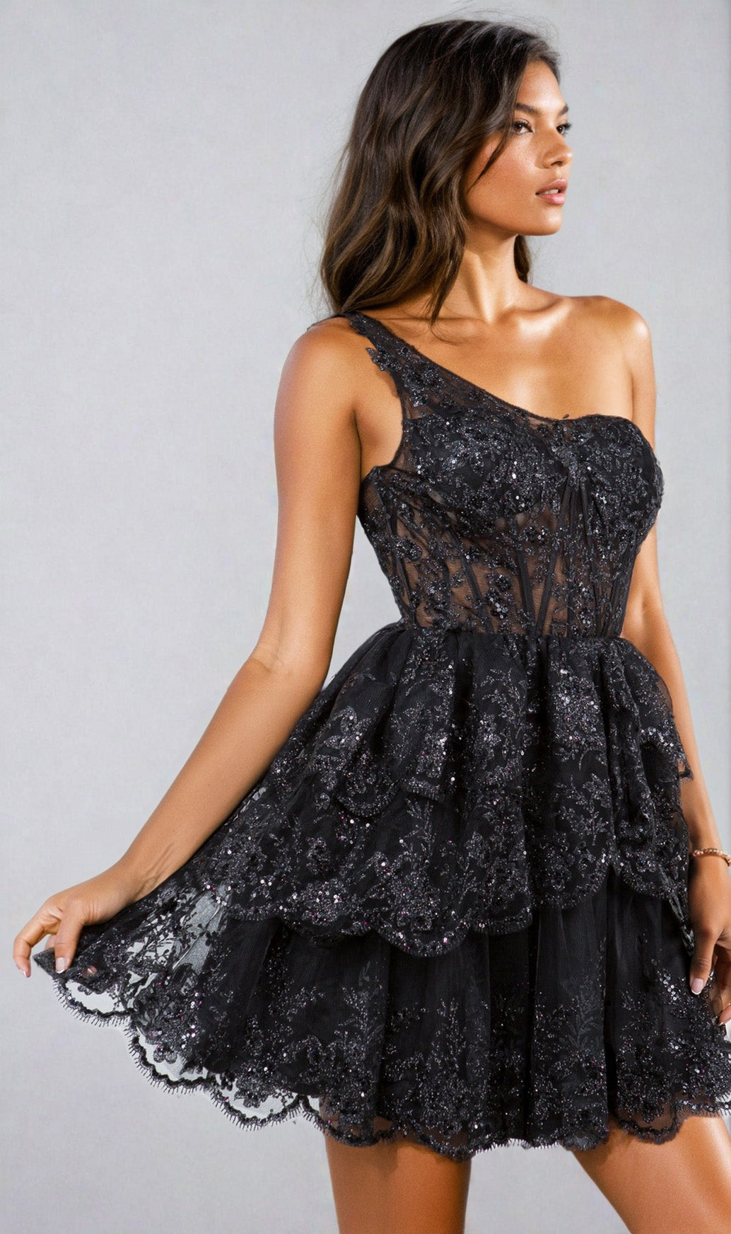 A woman models the One-Shoulder Short Tiered Homecoming Dress 5132J—a black, lace semi-formal dress with layered ruffles—while standing against a plain light background.