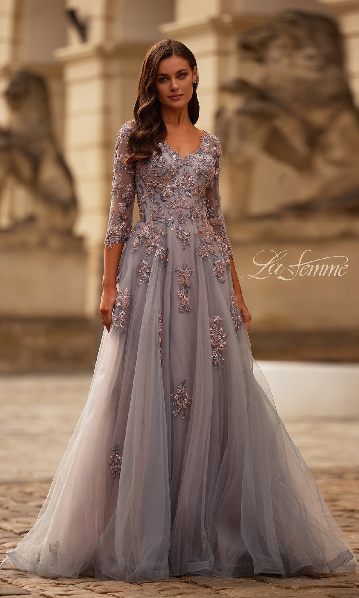 A woman stands outdoors in front of a stone building adorned with statues, wearing the Pink and Silver Long Formal Dress: La Femme 32605.