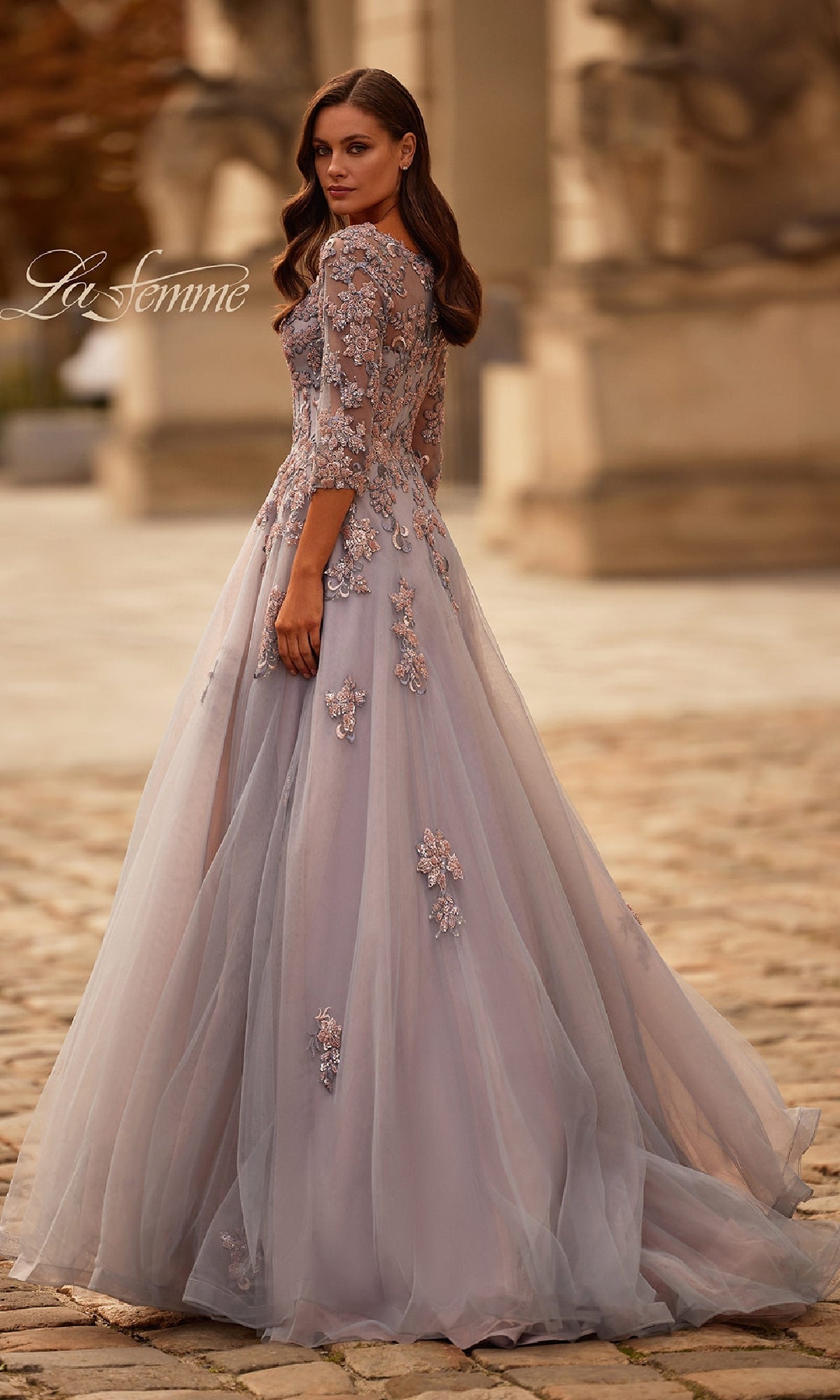 A woman in the La Femme 32605—a pink and silver long formal dress—stands outdoors on a stone pathway, looking over her shoulder.