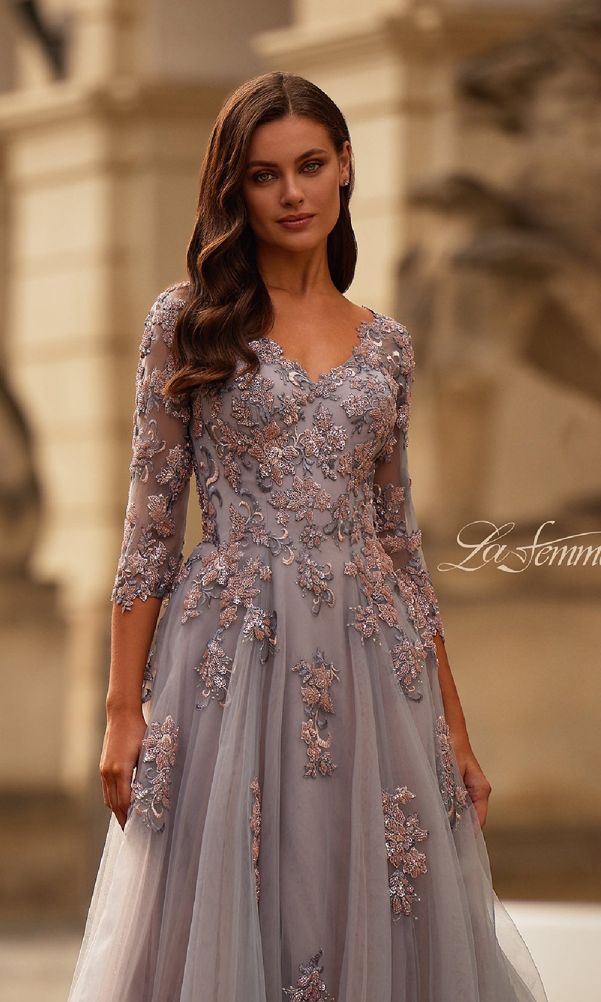 A woman stands outdoors in front of a stone building wearing the Pink and Silver Long Formal Dress: La Femme 32605, featuring three-quarter sleeves and floral embroidery.