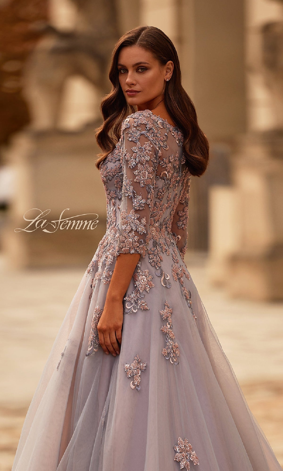 A woman stands outdoors, looking over her shoulder in the La Femme 32605, a pink and silver long formal dress with three-quarter sleeves and intricate lace detailing.