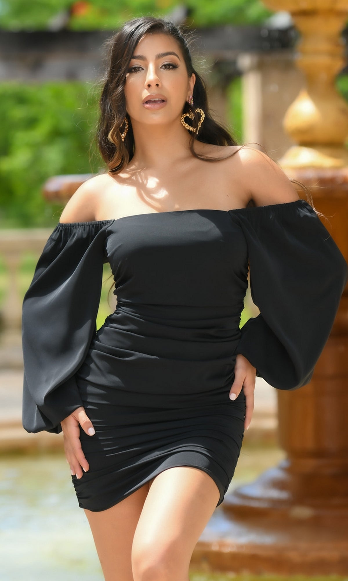 A woman stands outdoors in front of a fountain, wearing the Velvi Long Sleeve Short Graduation Dress Rhea with off-the-shoulder puffy sleeves.