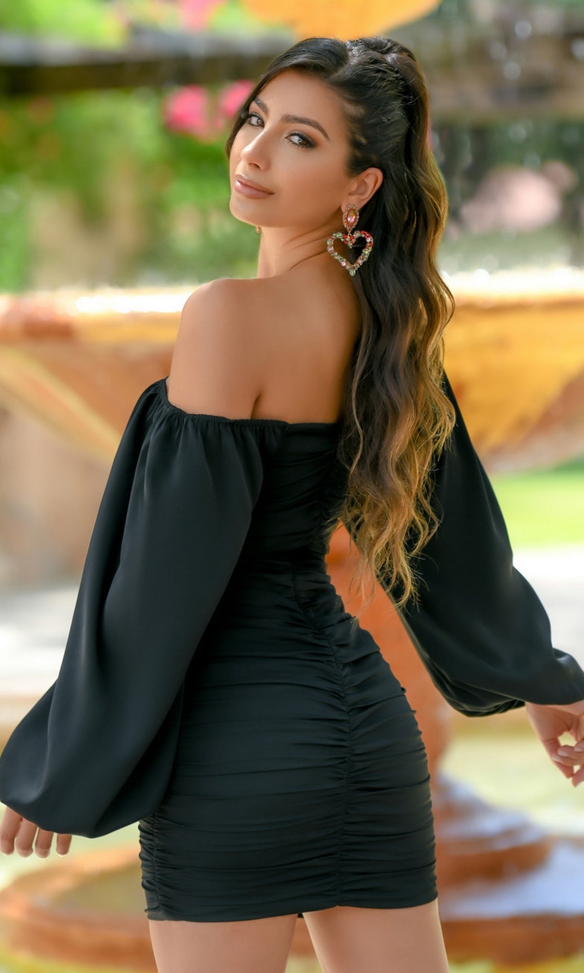 A woman with long wavy hair stands outdoors, looking over her shoulder while wearing the Velvi Long Sleeve Short Graduation Dress Rhea and heart-shaped earrings.