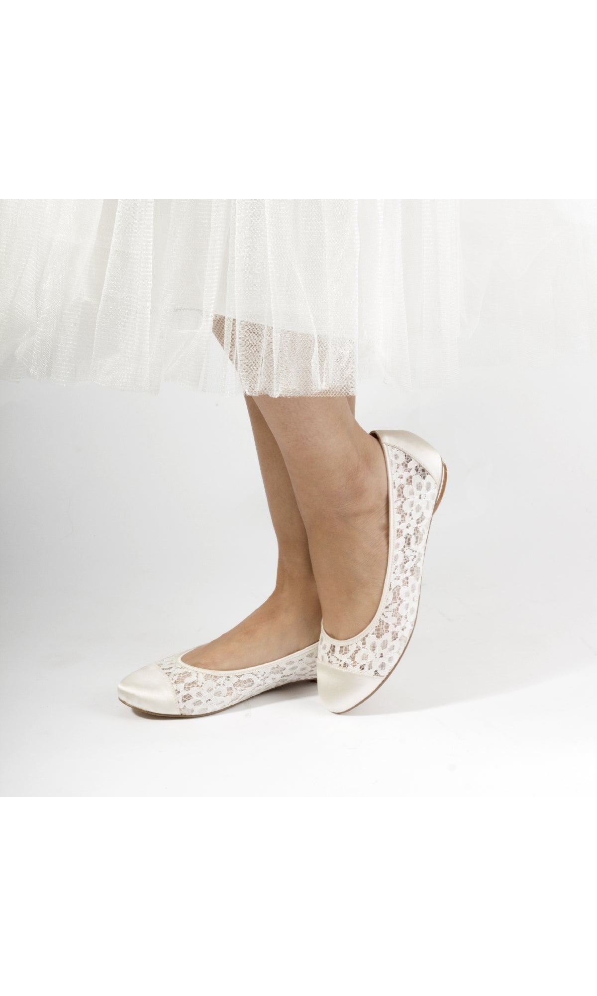 A person stands against a white background wearing a white tulle skirt and Sweetie Ivory Lace Prom Shoe Flats: Paradox London P1612—closed-toe slip-ons with embroidered floral patterns, an elegant prom choice.