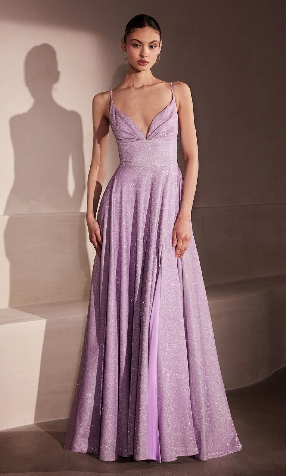 A woman stands indoors against a neutral background wearing the Amoris T214 Strappy-Back Glitter Prom Gown, a floor-length, sleeveless dress in shimmery lavender with a deep V-neckline and strappy-back design.