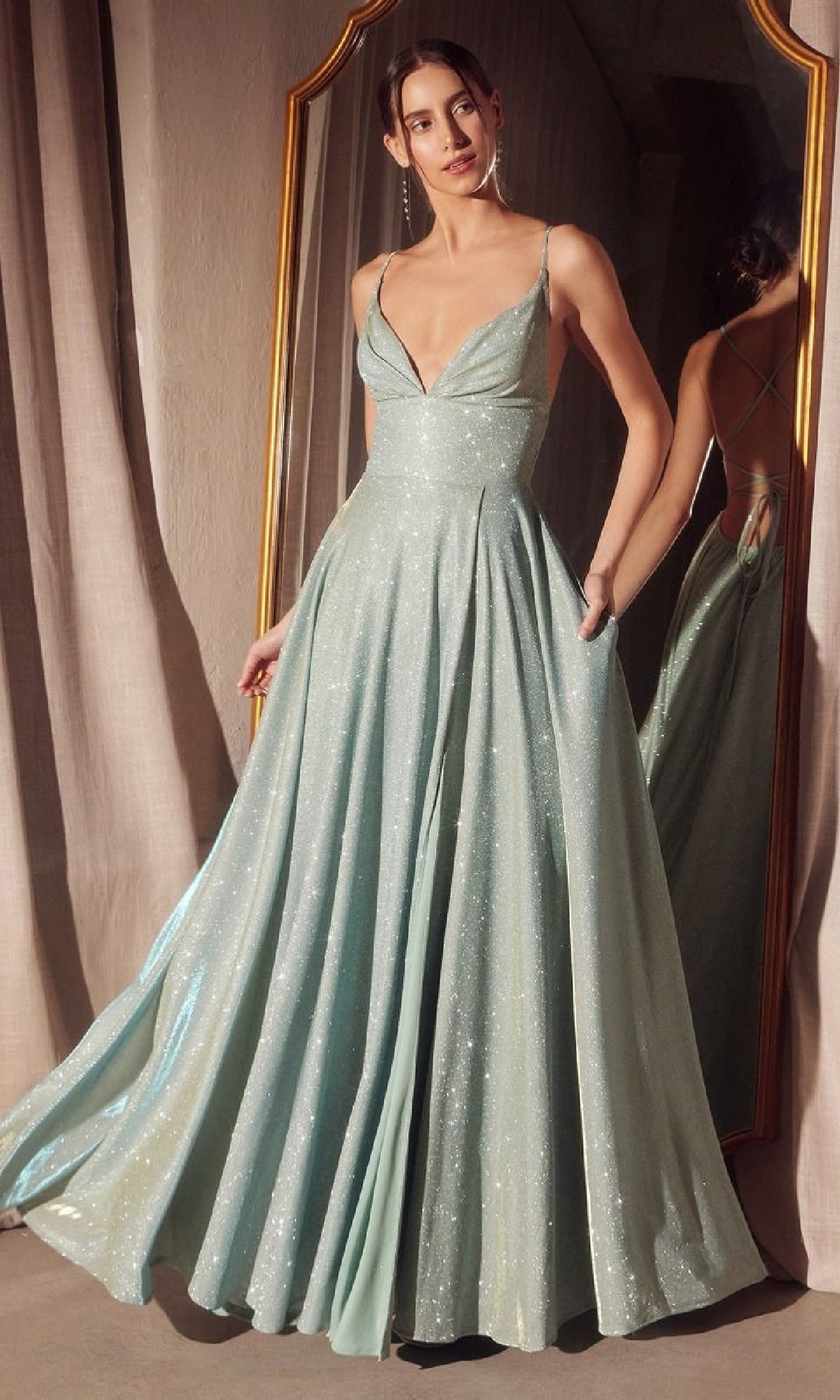 A woman in the Amoris T214 Strappy-Back Glitter Prom Gown, a shimmering light blue dress with thin straps, stands before a gold-framed mirror.