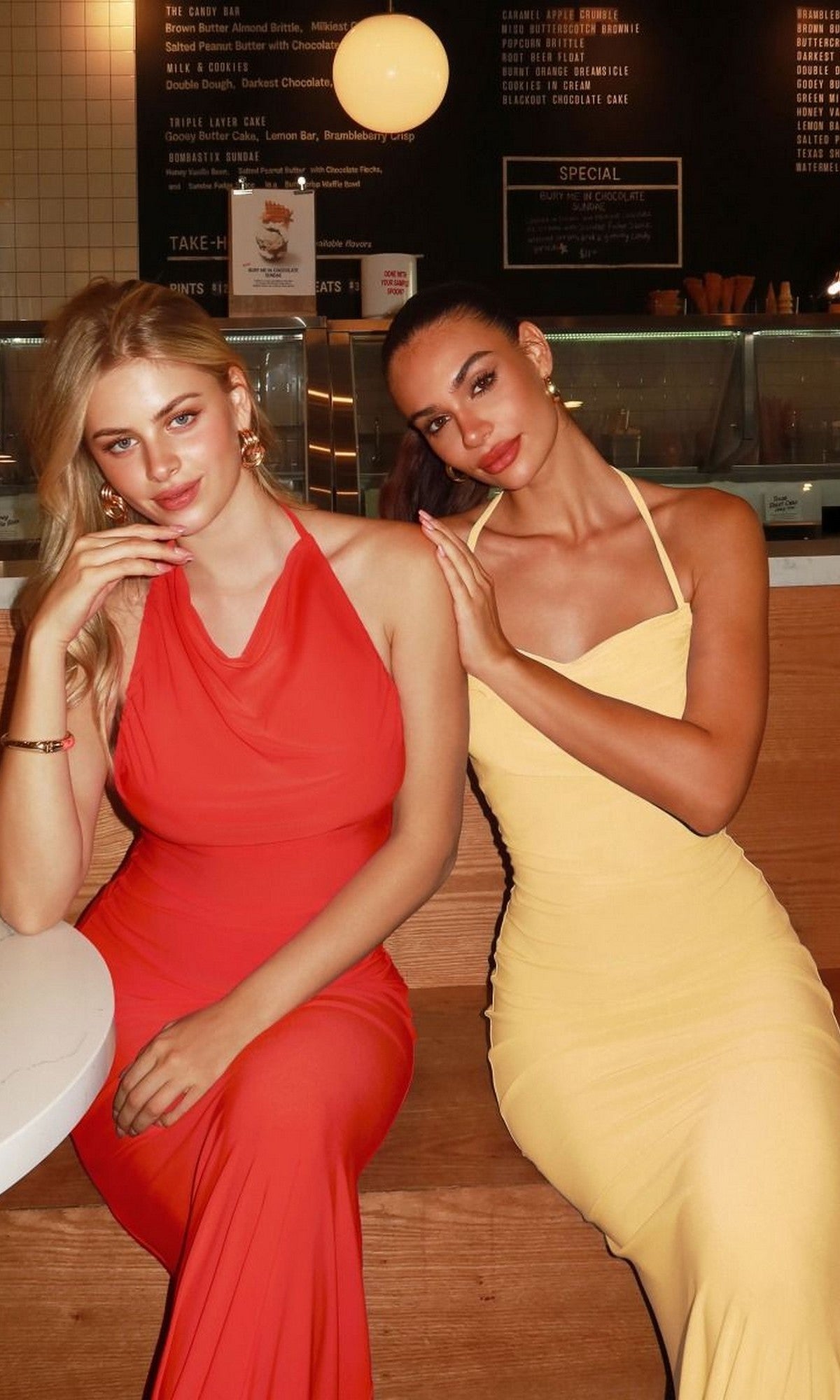 Two women sit side by side at a cafe table, one wearing a sleeveless red dress and the other in the Astee T506 long prom dress in light yellow; a menu is displayed on the wall behind them.