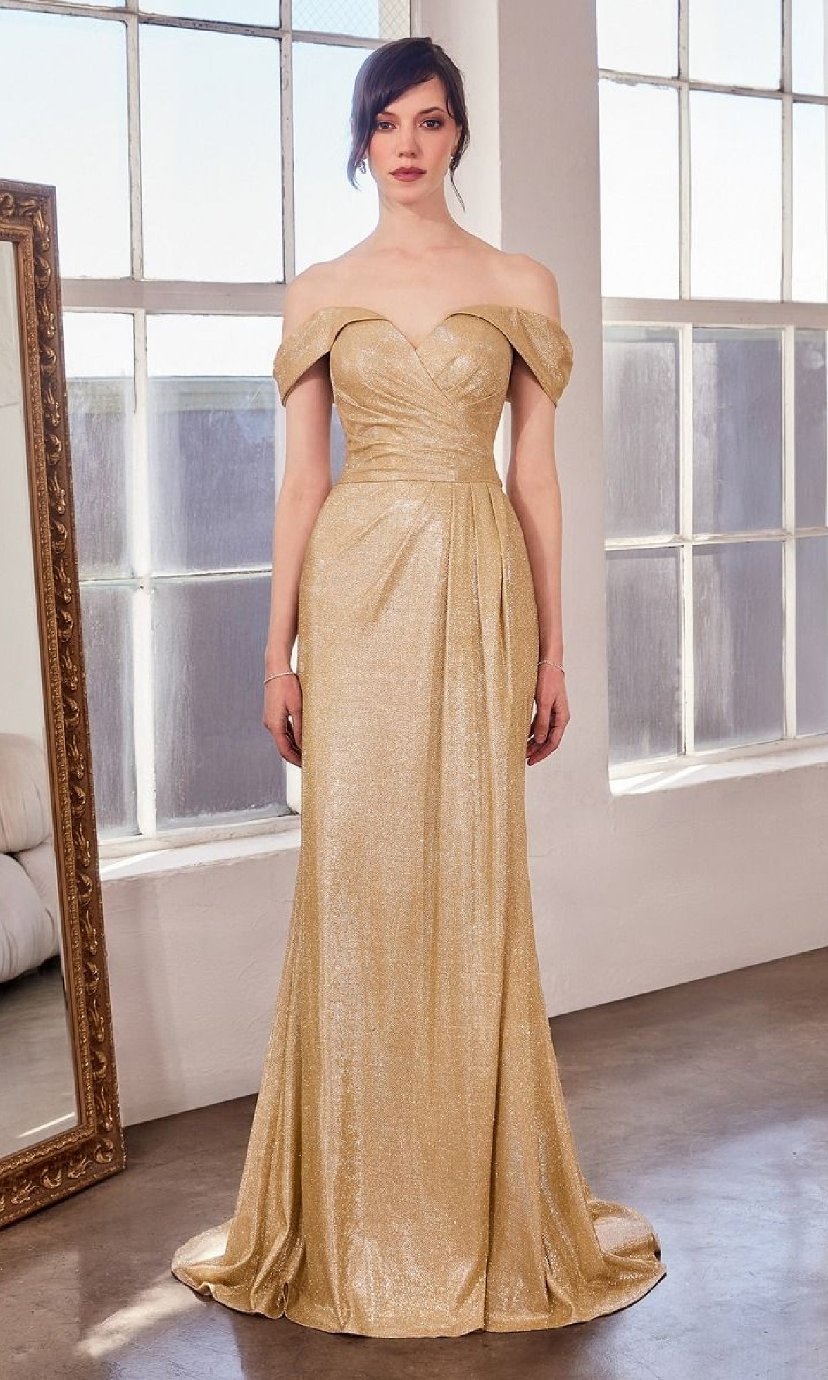 A woman stands indoors wearing the Off-the-Shoulder Glitter Prom Gown: Amoris T605, featuring a fitted bodice and floor-length gold shimmer. Large windows and a framed mirror are in the background.