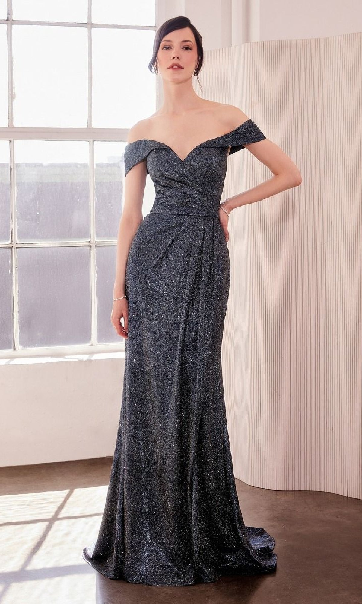 A woman in the Amoris T605 Off-the-Shoulder Glitter Prom Gown stands by a large window, her floor-length evening dress shimmering in dark blue indoor light.