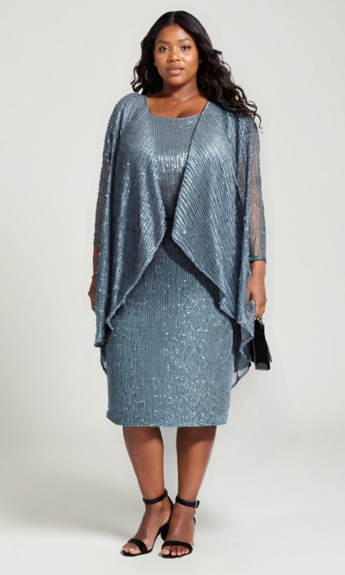 A woman models the Plus-Size Two-Piece Party Dress: Amoris T9001, featuring a silver-blue sequin knee-length dress with a matching sheer overlay and black ankle-strap heels, against a plain background.