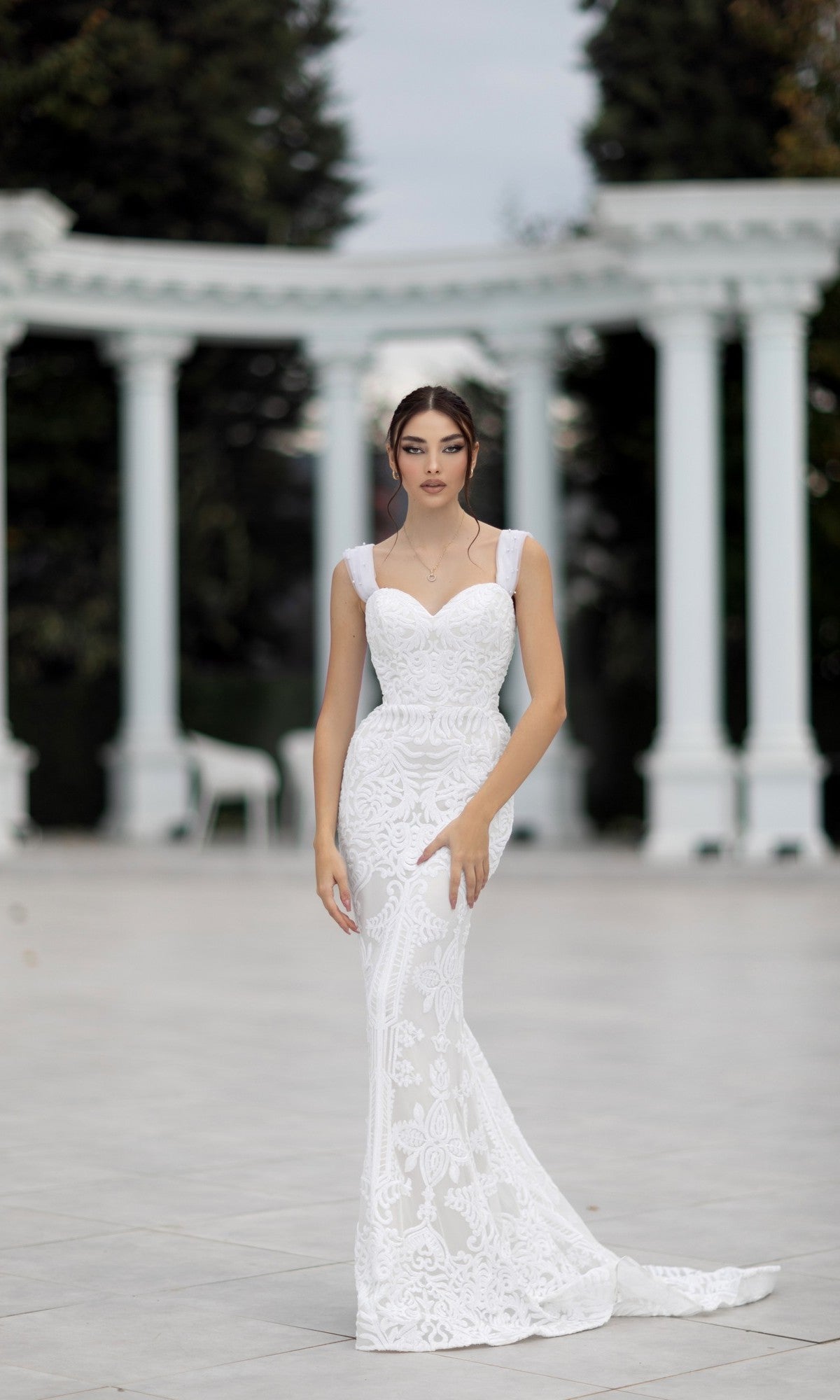 A woman wearing the Off-Shoulder White Bridal Gown: Tina Holly TW103 stands outdoors before white columns and lush greenery.