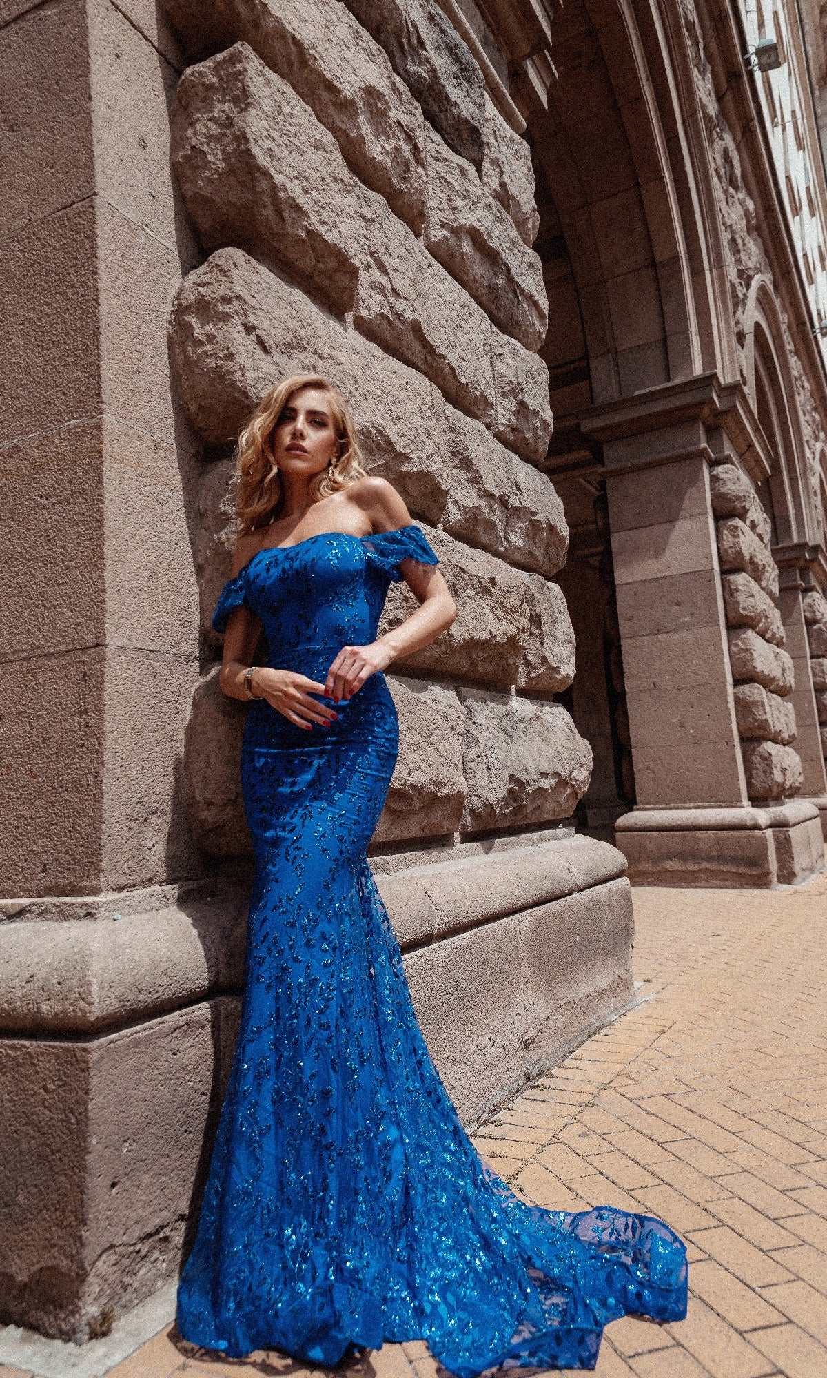 Woman wearing the Long Glitter-Lace Prom Dress: Tina Holly TW024, featuring off-the-shoulder sequin details, stands by a stone building with elegant arched architecture.