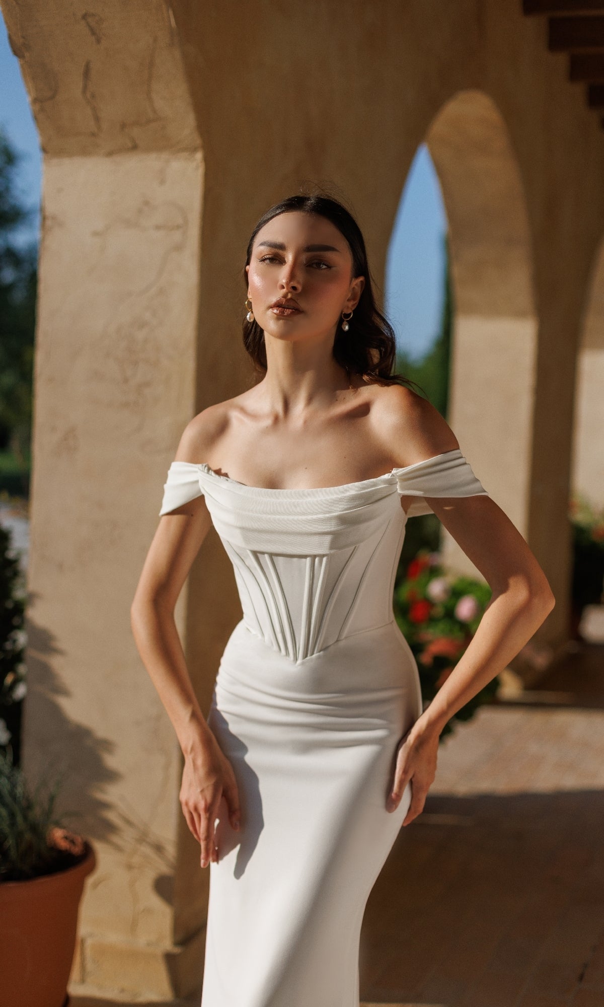 A woman stands outdoors under an archway, wearing the Off-White Long Bridal Gown: Tina Holly TW485, featuring an off-the-shoulder design with a fitted bodice and minimal jewelry.