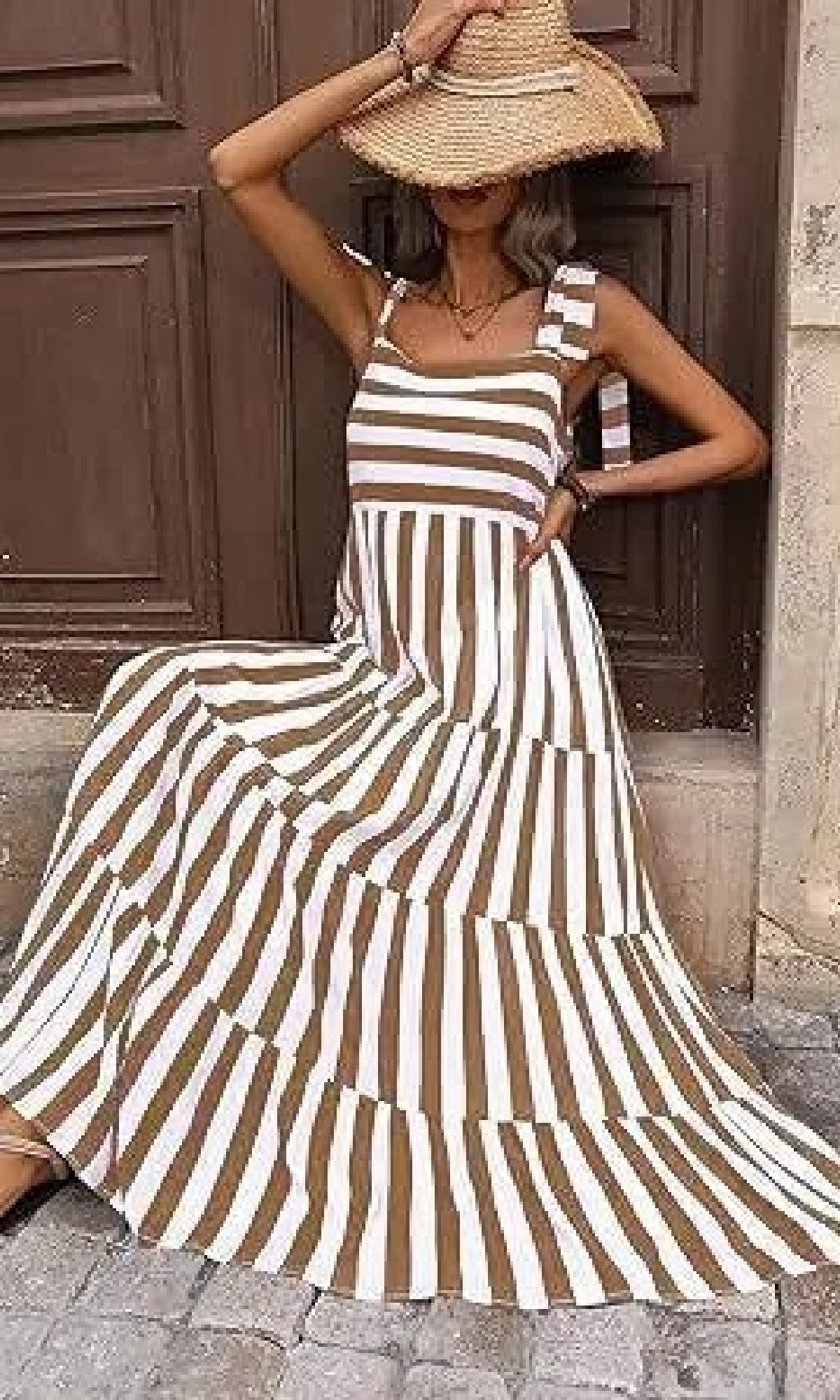 A woman poses on stone pavement in front of a wooden door, wearing the Striped Casual Relaxed-Fit Maxi Dress and a wide-brimmed straw hat, with one arm raised to her head.