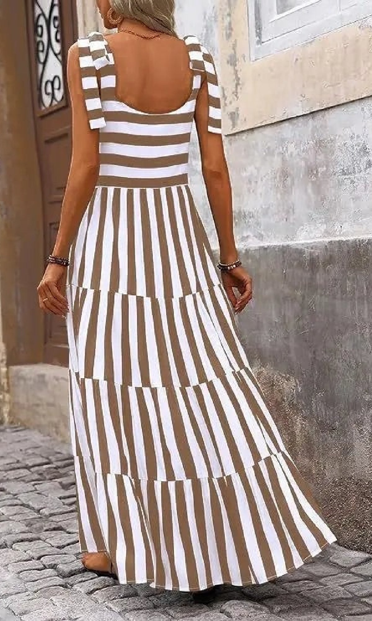A woman in a Striped Casual Relaxed-Fit Maxi Dress strolls along a cobblestone street beside a weathered wall.