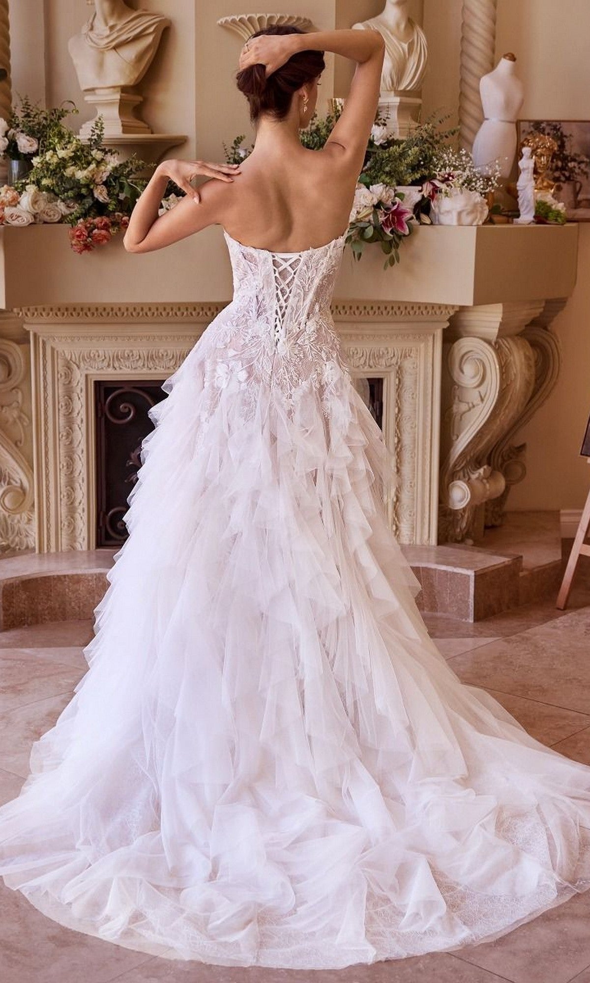 A woman in the Andrea and Leo WL016 long wedding dress, with a strapless lace-applique corset bodice and ruffle tulle a-line skirt, stands facing away in an ornate room with floral arrangements and classical statues.