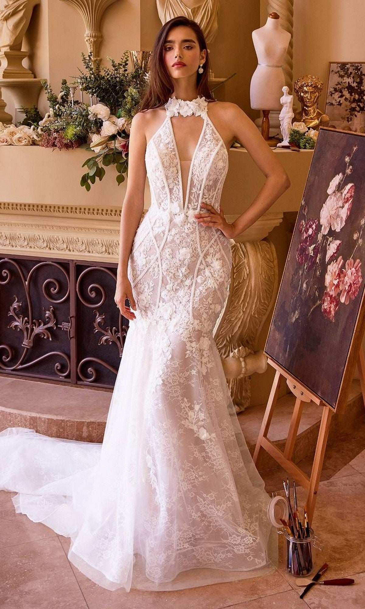 A woman in the Andrea and Leo WL019 long fitted white lace mermaid wedding dress stands indoors near a decorated fireplace and a floral painting on an easel.