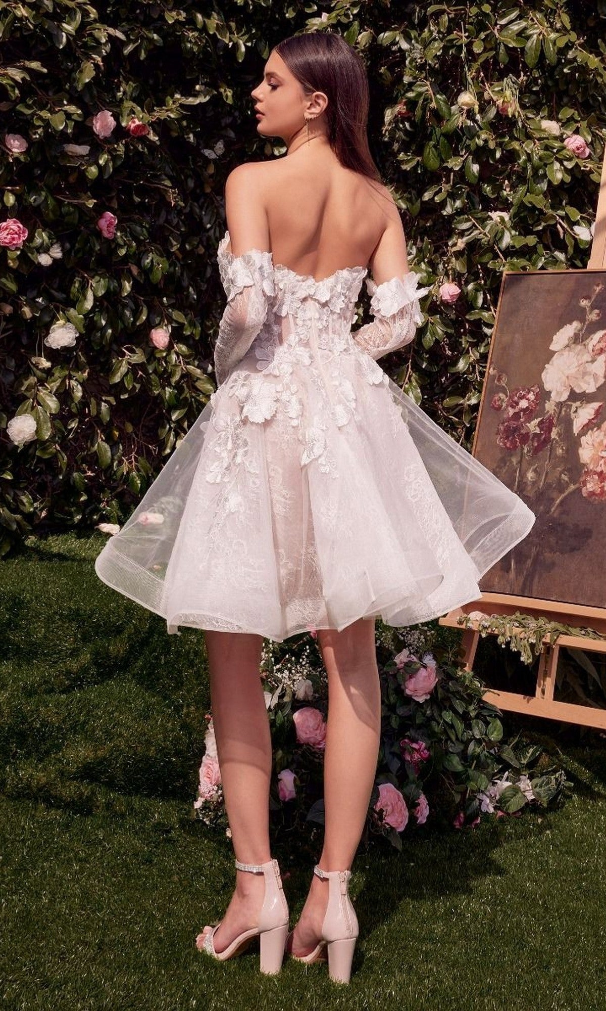 A woman stands in a garden wearing the Short White Party Dress: Andrea and Leo WL020, featuring floral appliques and sheer sleeves. She faces away from the camera amid lush greenery and a floral painting, creating an enchanting scene.