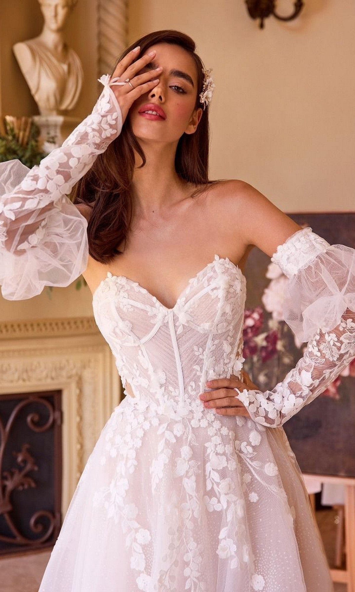 A woman wearing the Andrea and Leo WL032 long wedding dress with a strapless sweetheart neckline and floral lace details poses indoors, her gloved hand partially covering her face.