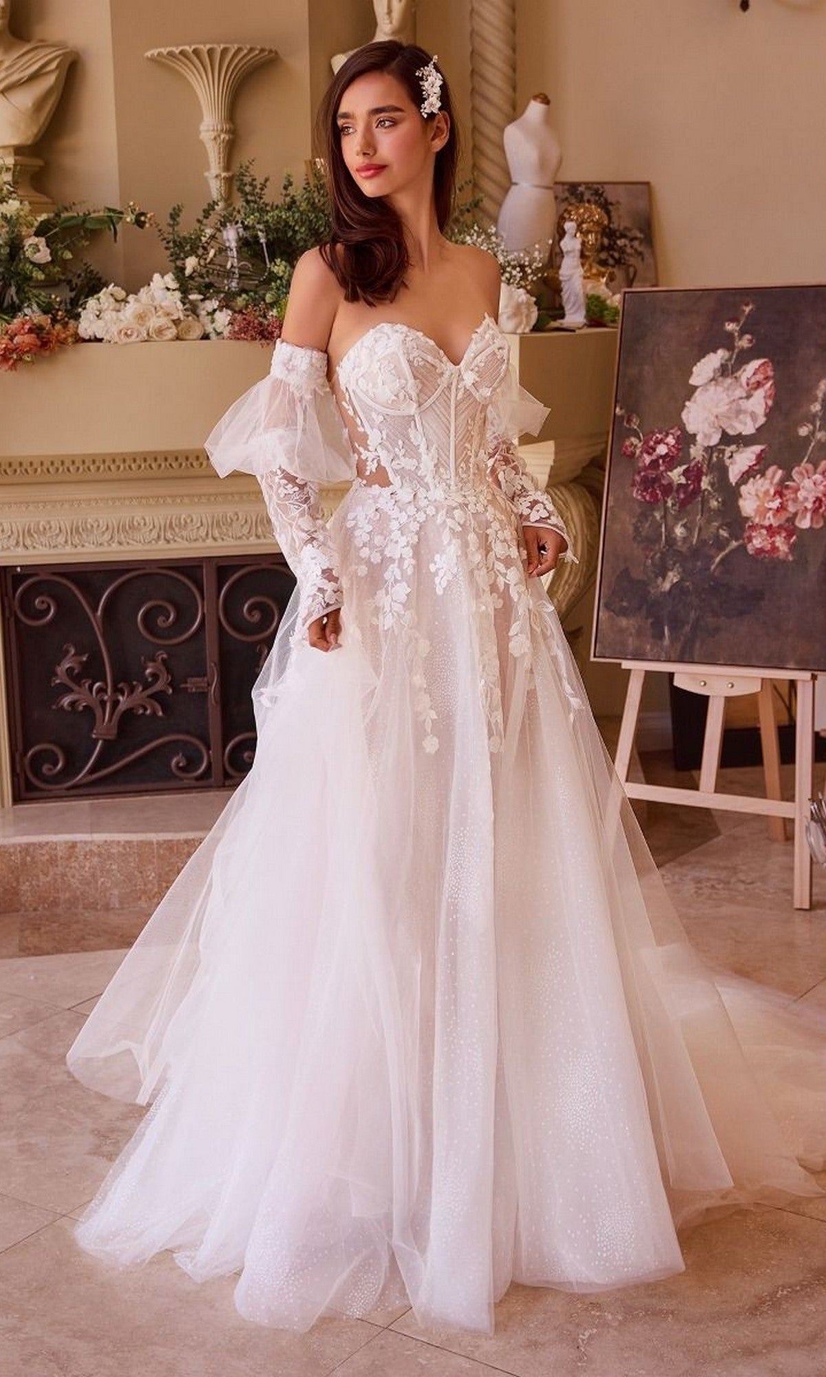 A woman wears the Long Wedding Dress: Andrea and Leo WL032, featuring a strapless bodice and an a-line skirt, as she stands indoors near a decorated mantel and a floral painting on an easel.
