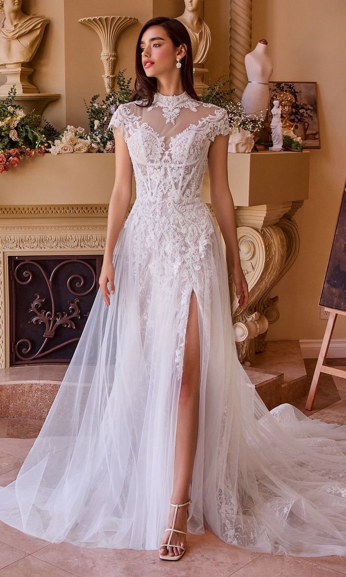 A woman poses indoors in front of a decorated fireplace, wearing the Andrea and Leo WL035 long wedding dress—a sheer lace gown with cap sleeves, tulle skirt, and thigh-high slit.