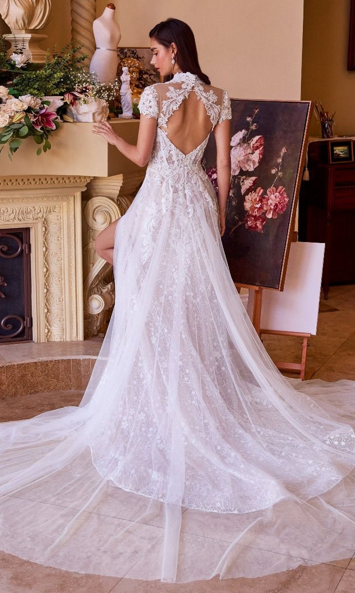 A woman wears the Long Wedding Dress: Andrea and Leo WL035, featuring a long lace train, as she stands indoors. A painting, floral arrangement, and dress form create an elegant backdrop for her stunning bridal look.