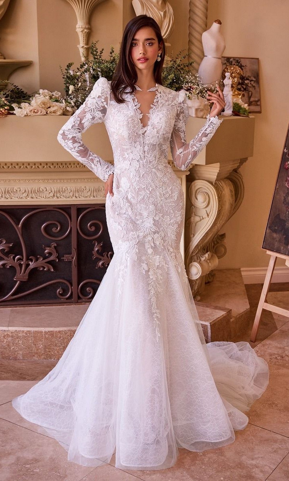 A woman stands indoors wearing the Andrea and Leo WL036 long wedding dress, featuring lace fabric, a mermaid silhouette, long sleeves, a deep V neckline, and exquisite floral detailing.
