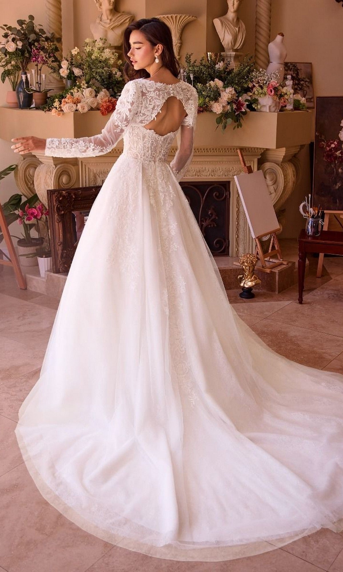 A woman poses indoors in front of a flower-adorned fireplace, wearing the Andrea and Leo WL037 long wedding dress featuring lace sleeves, a keyhole back, and a lace bodice.