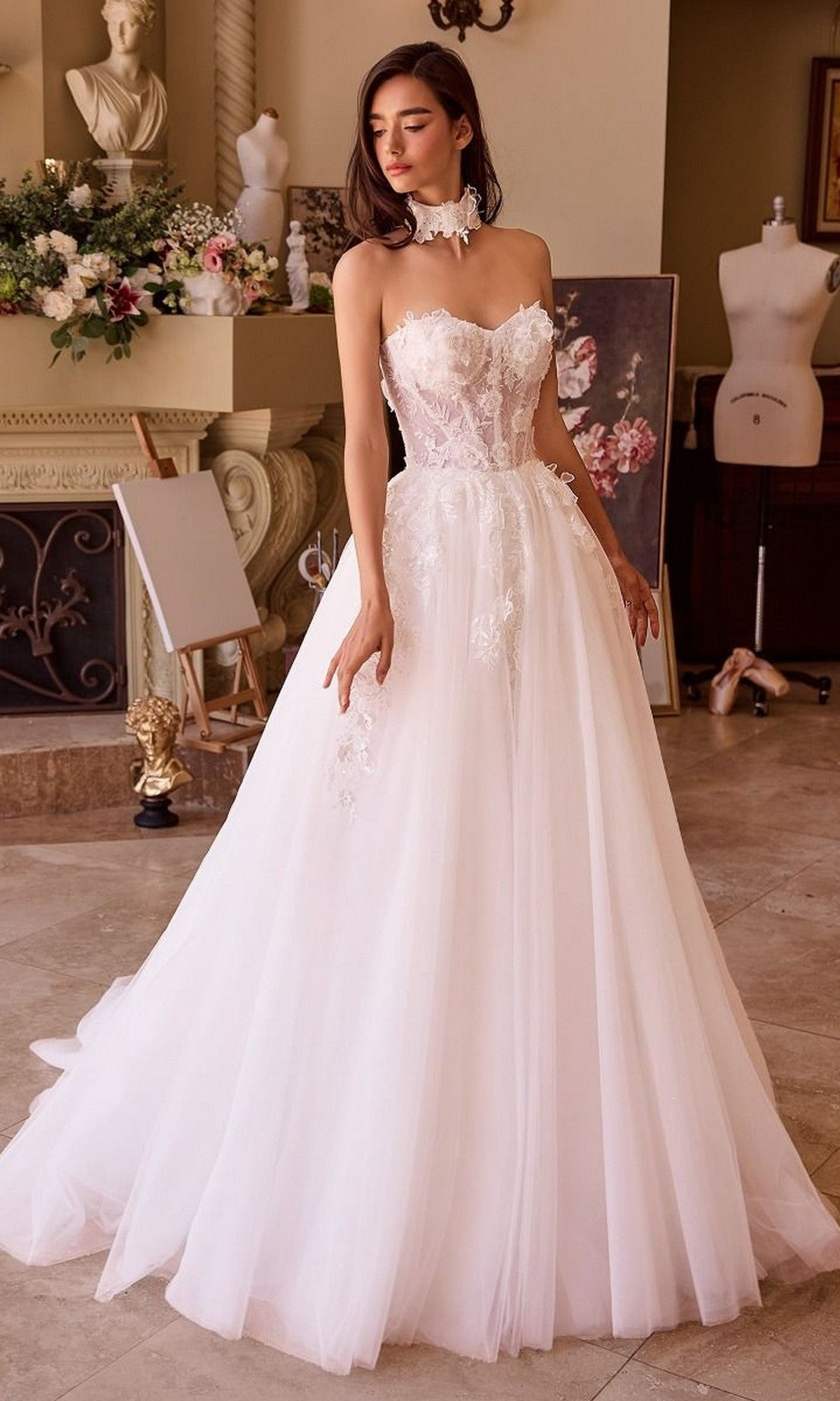 A woman wears the Andrea and Leo WL041 long wedding dress, featuring a strapless floral lace bodice and elegant A-line tulle skirt, standing indoors by floral décor and a dress form.