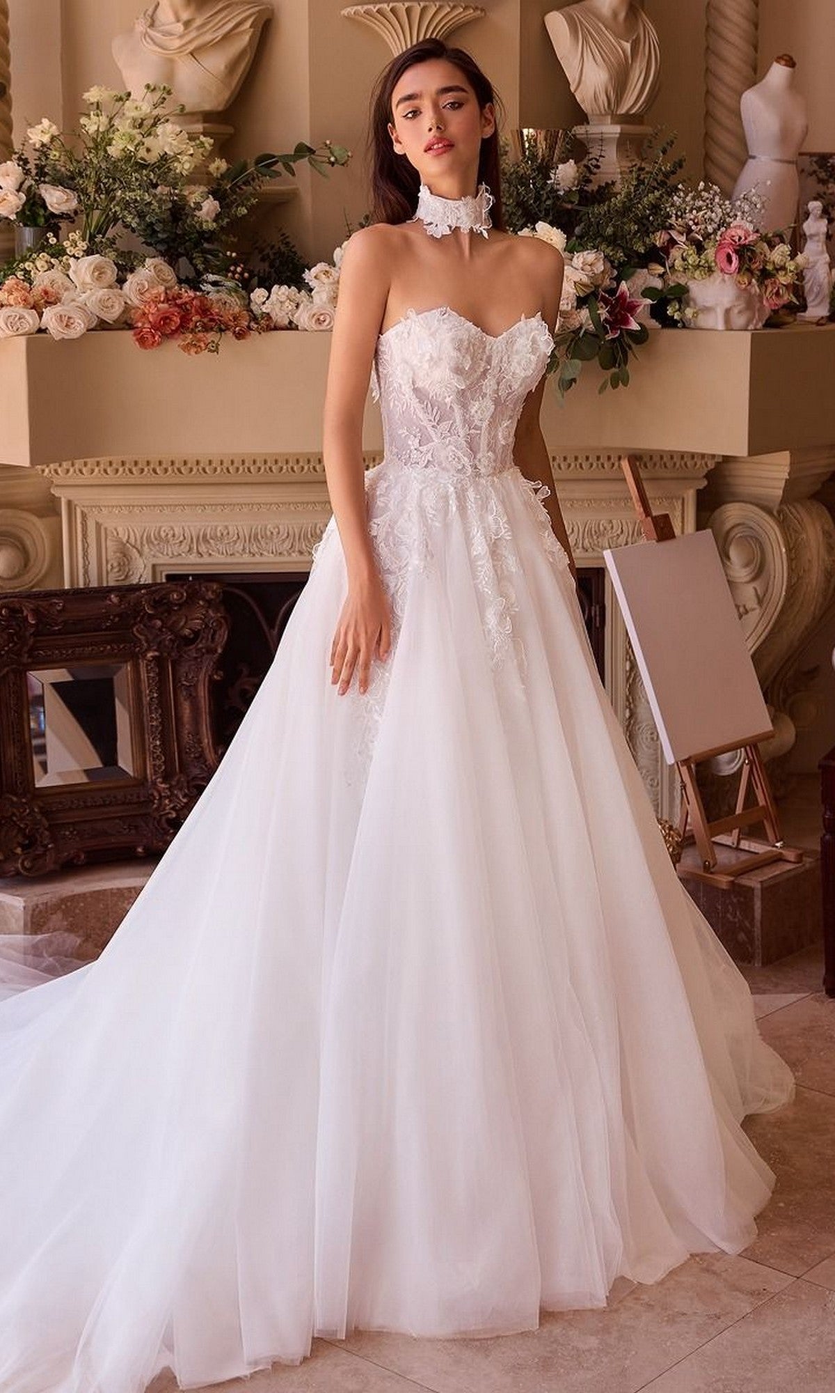 A woman stands indoors wearing the Andrea and Leo WL041 long wedding dress, featuring a strapless floral lace bodice and full A-line tulle skirt, posed in front of a flower-adorned mantel.