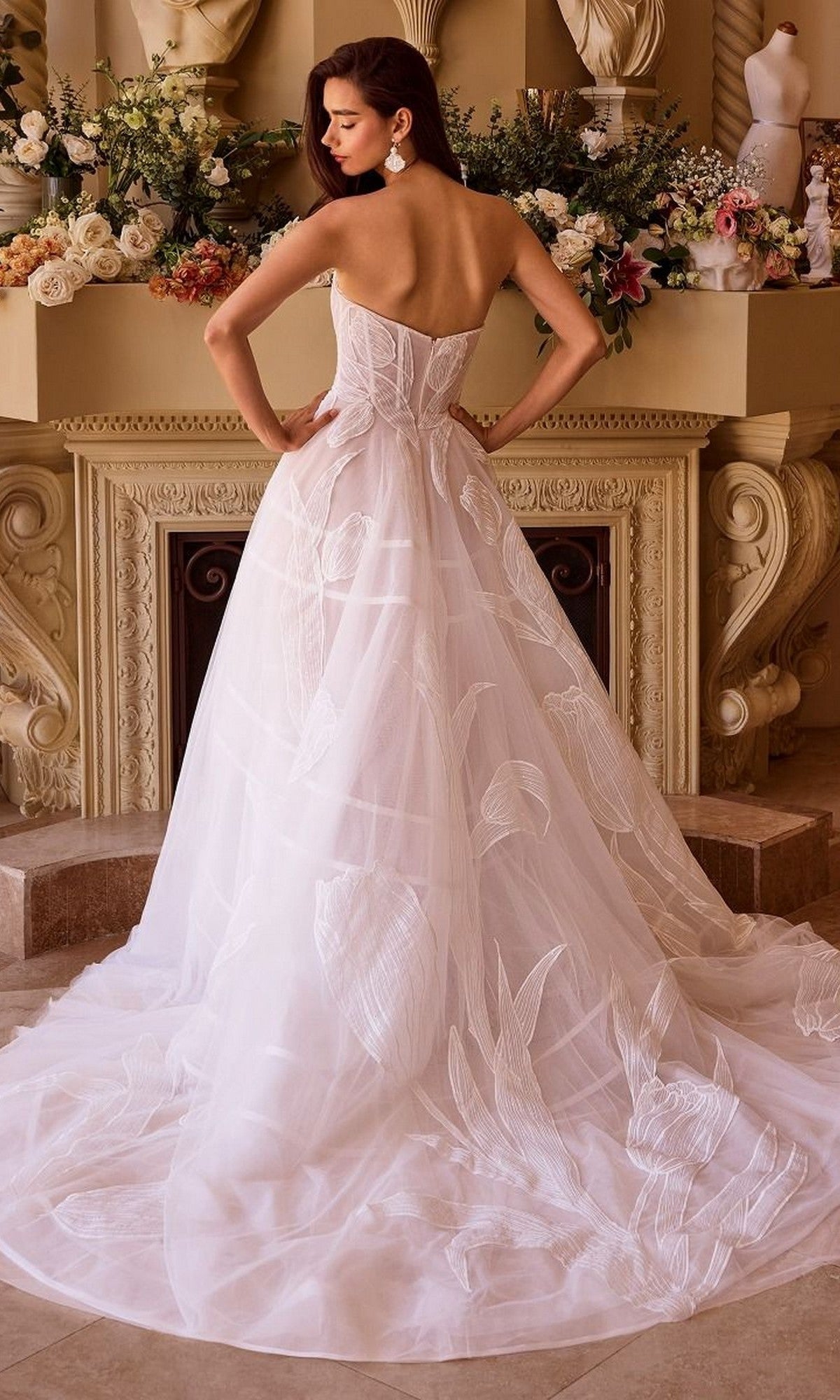 A woman wearing the Andrea and Leo WL043 long wedding dress with a strapless sweetheart neckline and sheer tulle A-line skirt with leaf patterns stands indoors before a decorated fireplace.